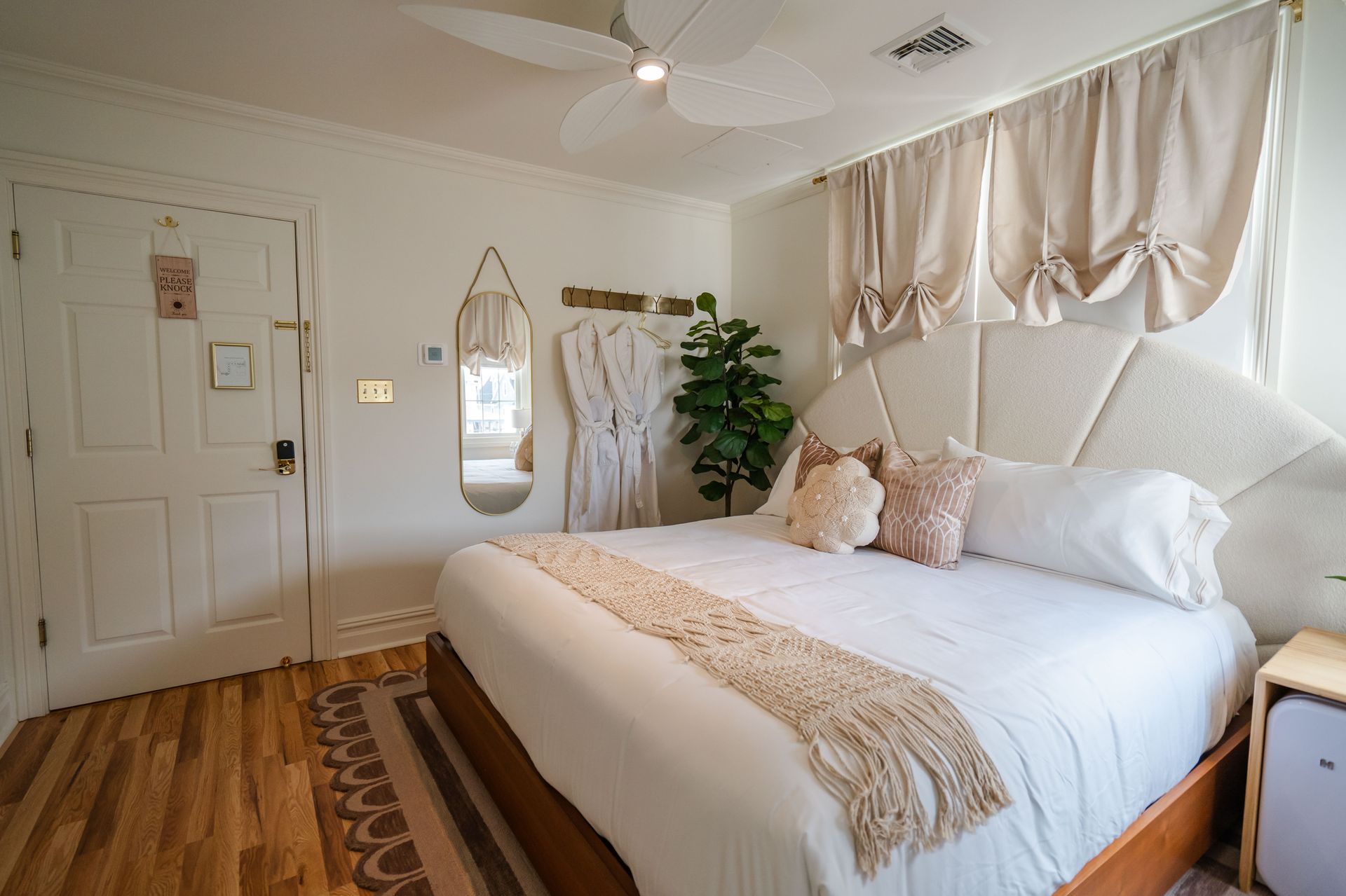 Cozy bedroom with bed, beige headboard, cream curtains, wooden floor, and a tall plant.