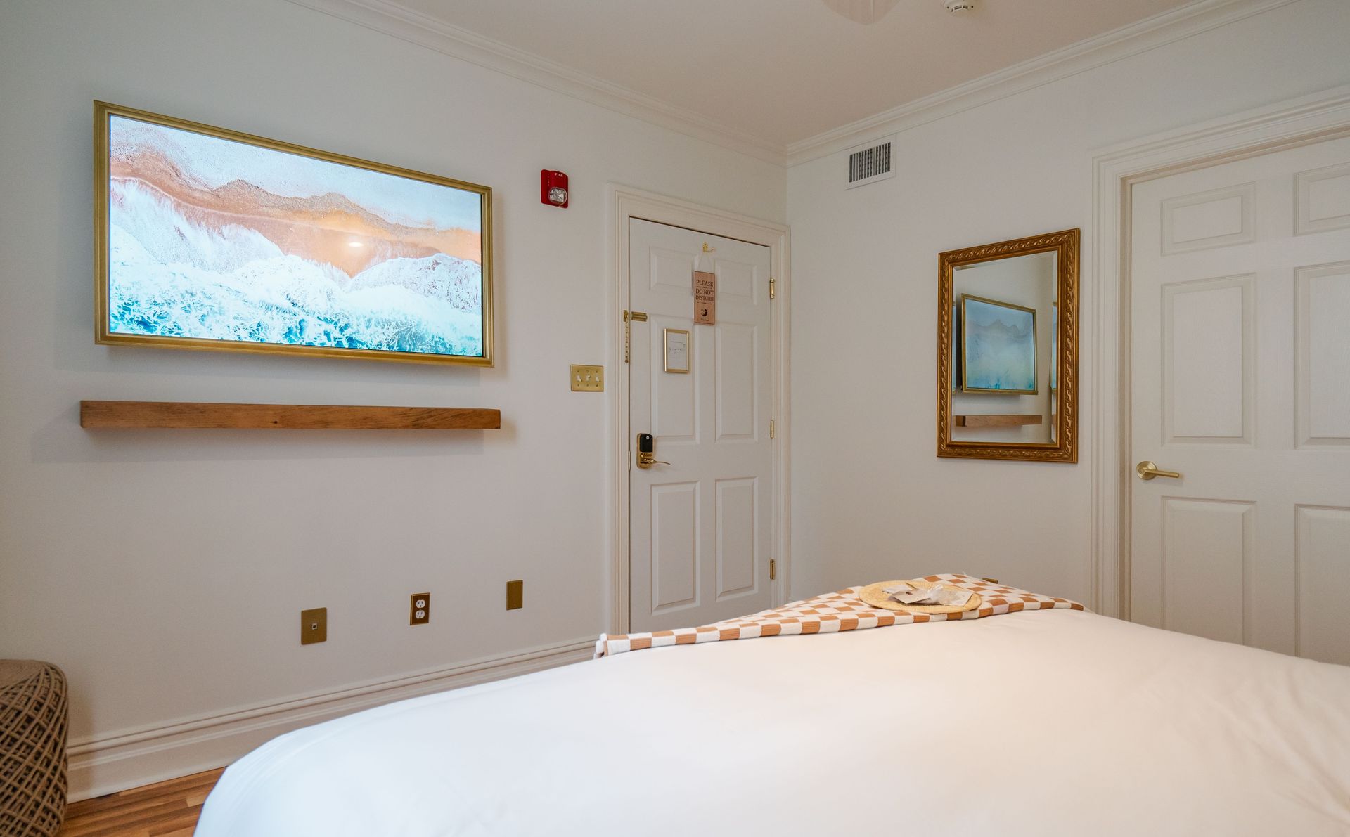 Hotel bedroom with a bed, TV, mirror, and door against a white wall.
