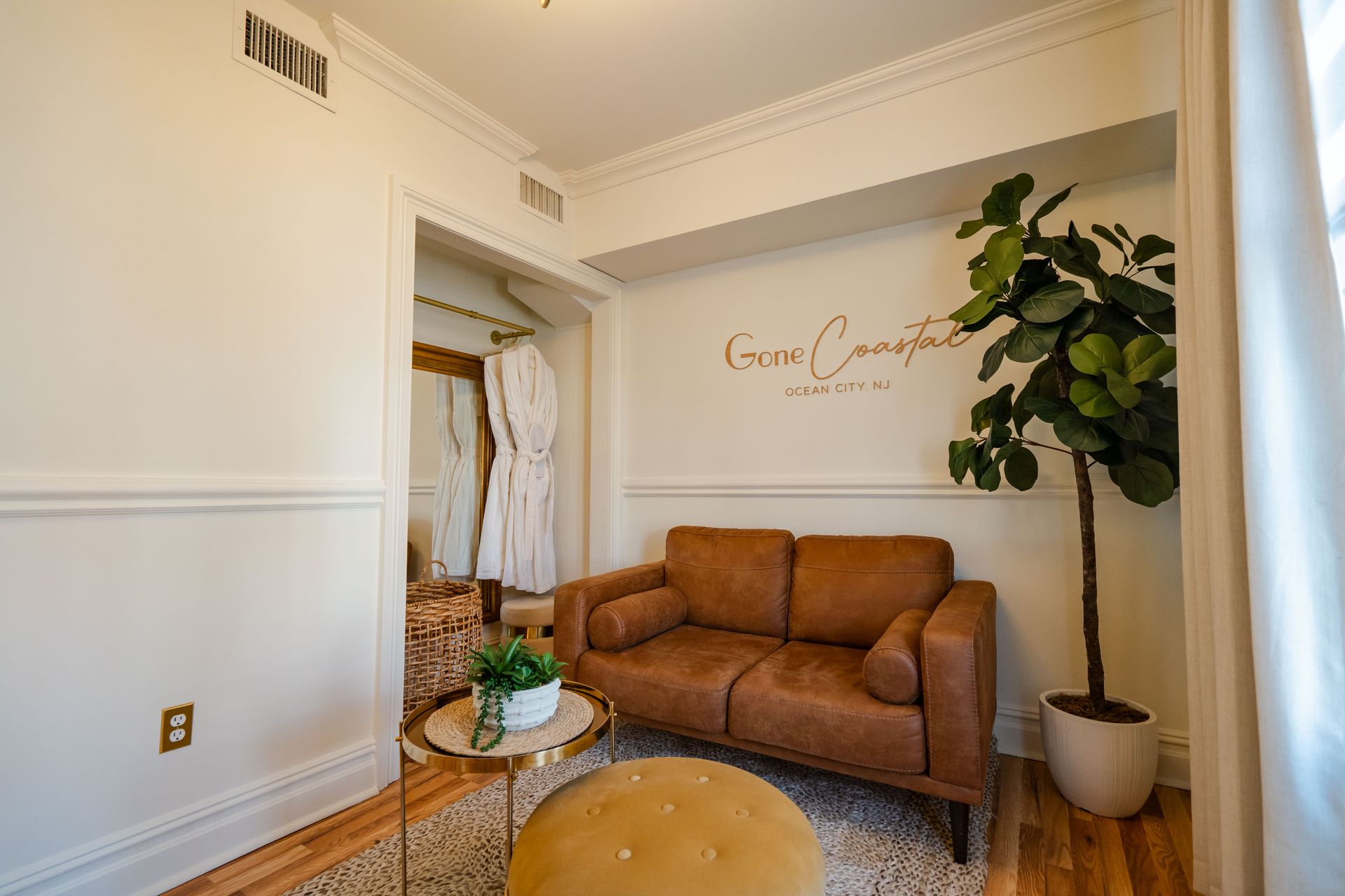 A cozy room with a brown couch, plant, and gold accents. A dressing area is visible in the background.