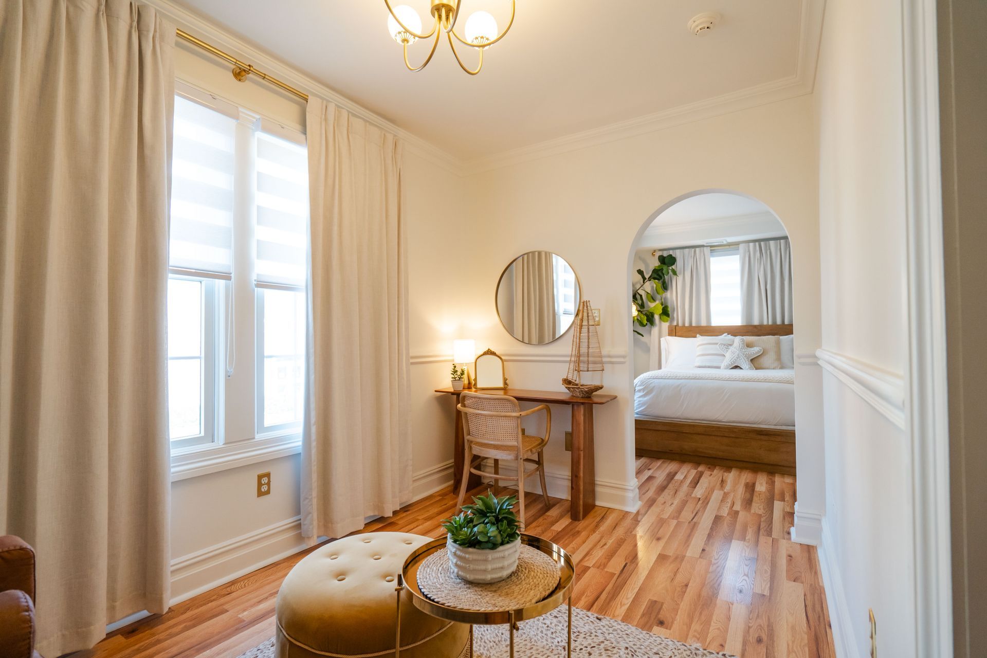 Bright, cozy room with a vanity, round mirror, and an arched doorway leading to a bedroom with a bed.
