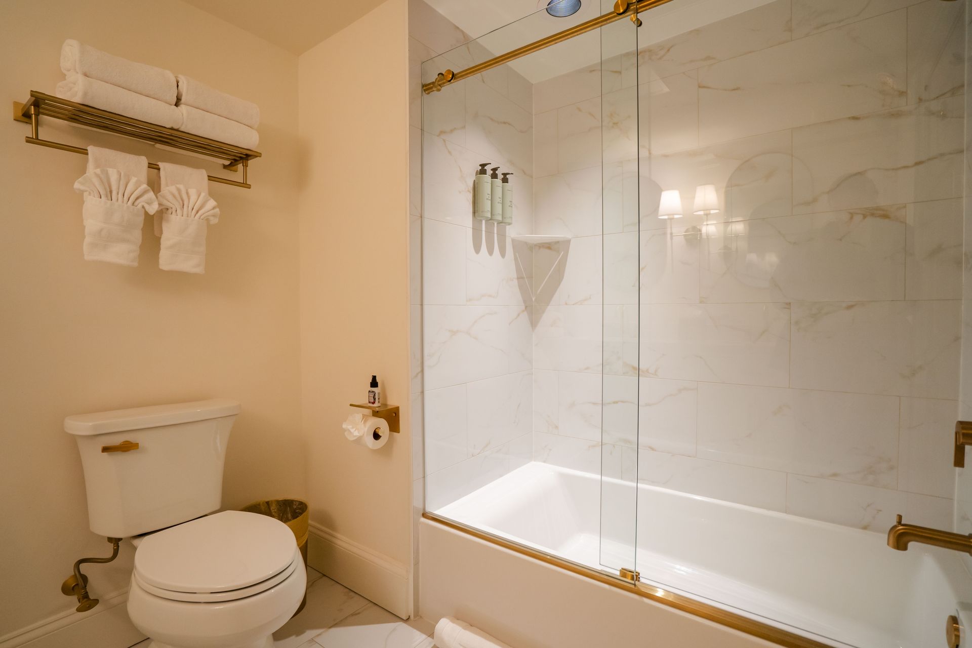 Bathroom with toilet, tub/shower with glass door, towels on rack. Beige walls, gold fixtures.