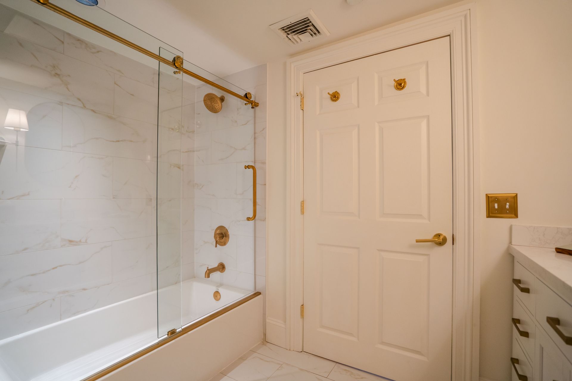 Bathroom with white marble tile, gold fixtures, glass shower door, and a closed white door.