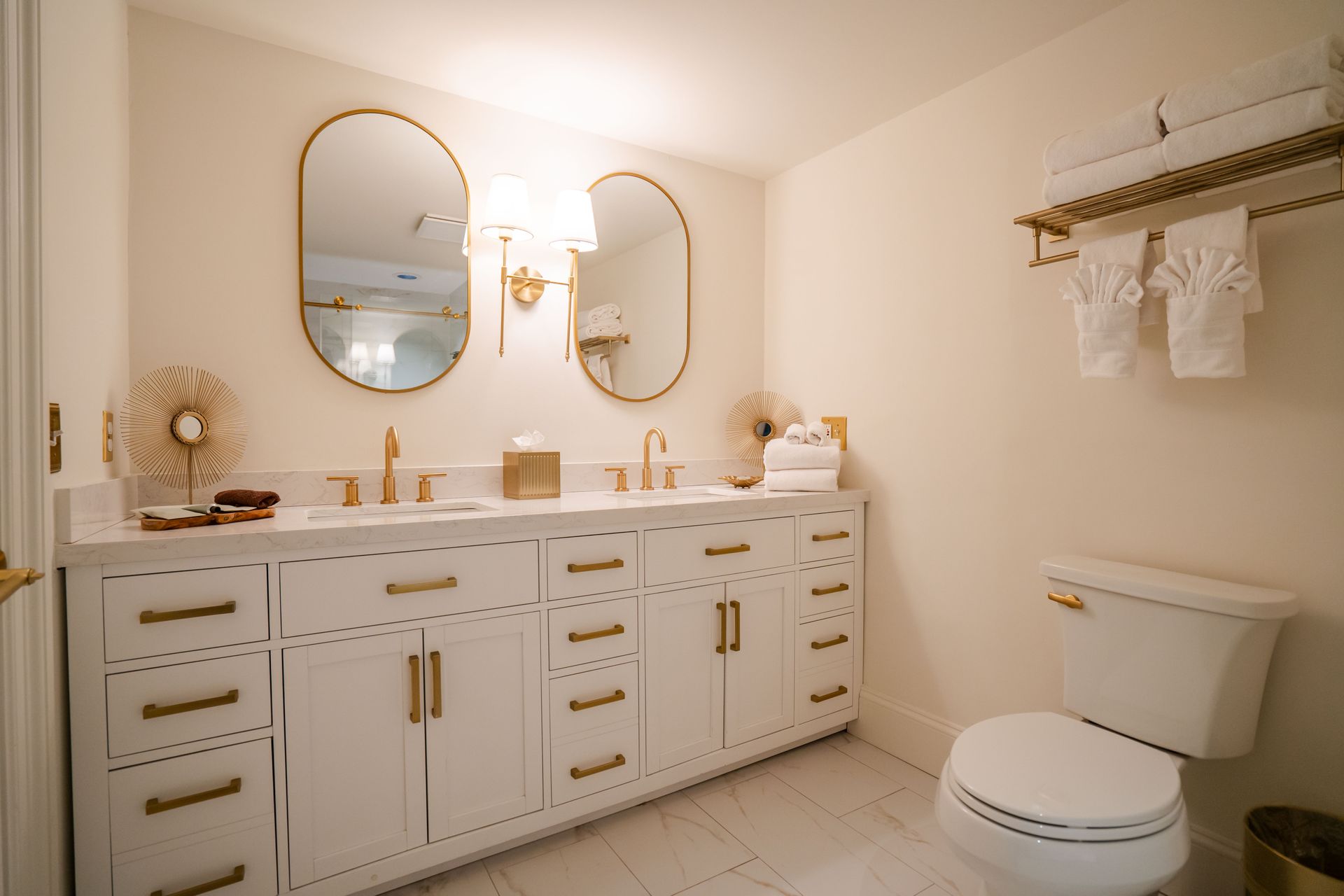 Elegant white bathroom with gold accents, double vanity, oval mirrors, and a towel rack.