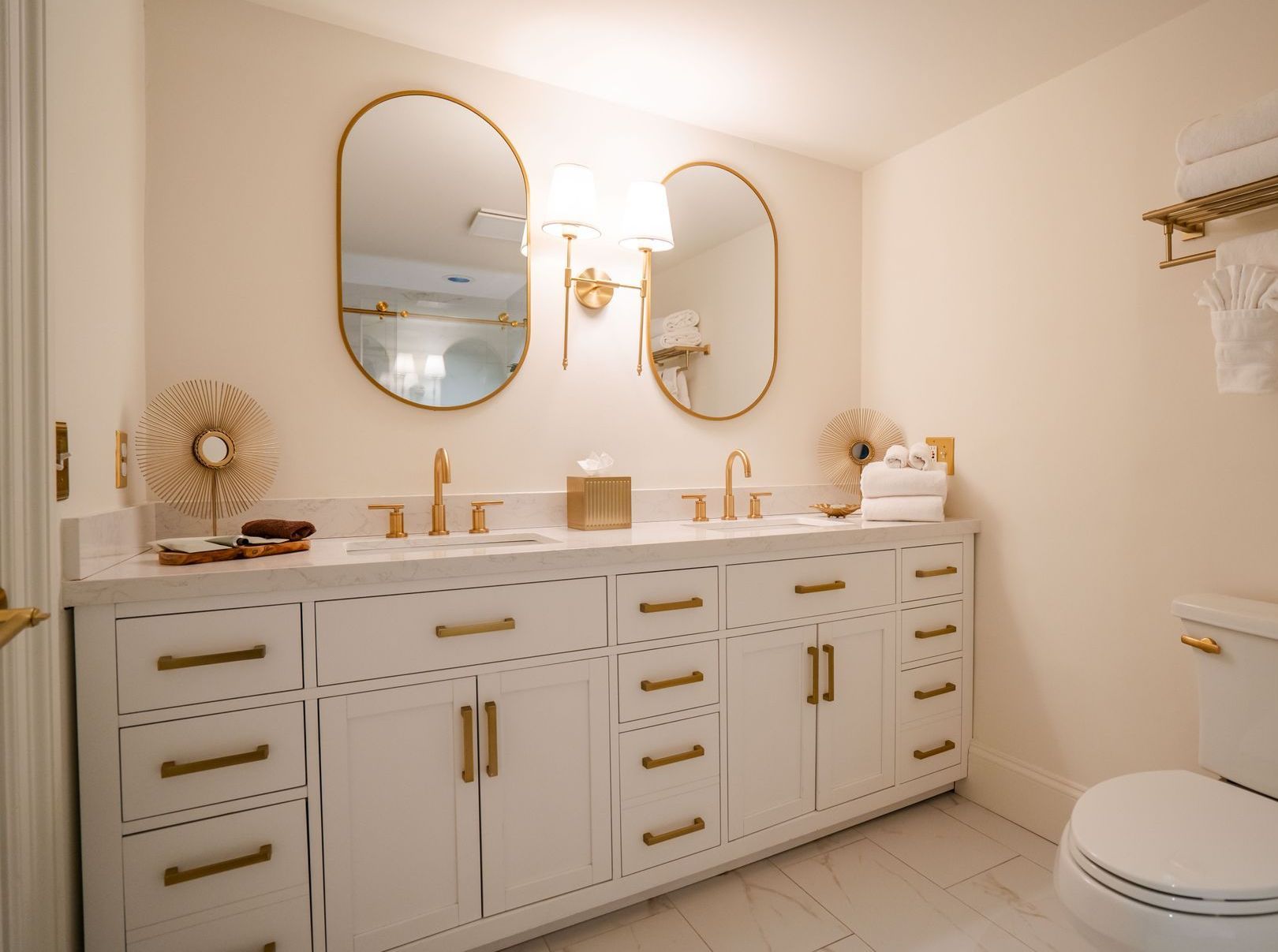 Bathroom with white cabinets, gold fixtures, oval mirrors, and a white toilet.