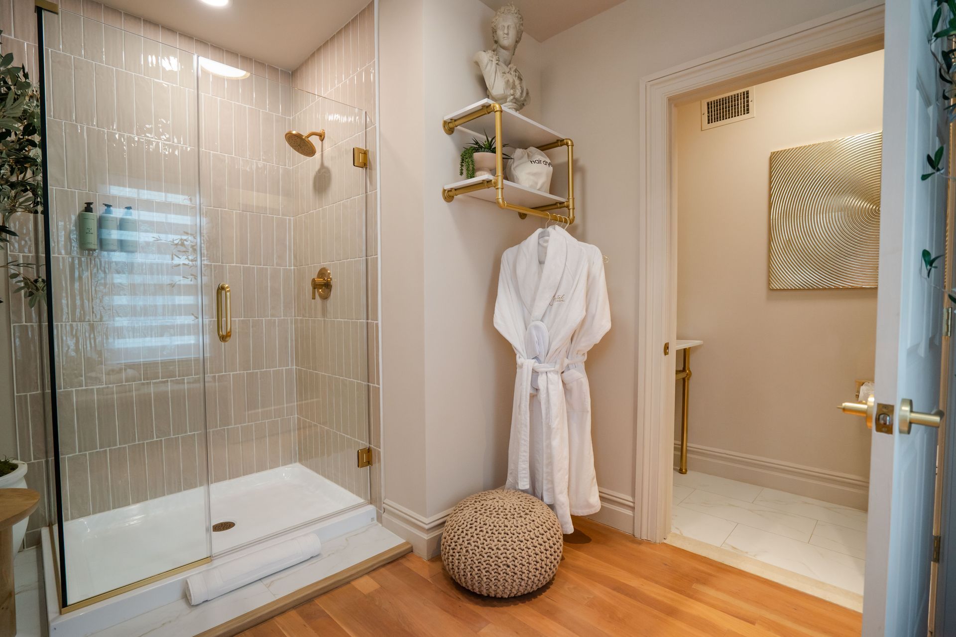 Elegant bathroom with glass shower, gold fixtures, robe, wooden floor, and decorative shelf.