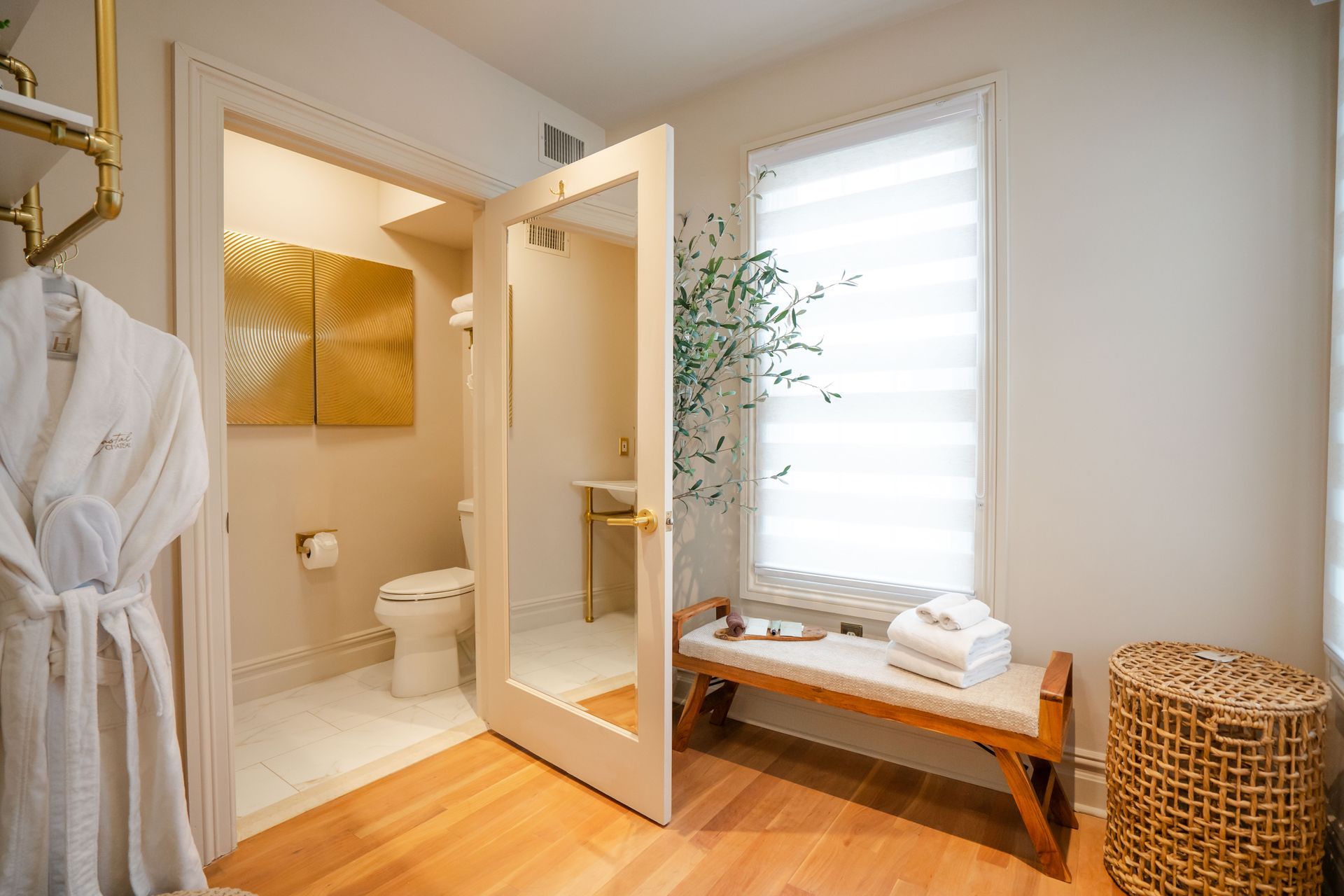 Spa-like room with an open doorway to a bathroom. White walls, gold accents, and a bench with towels.