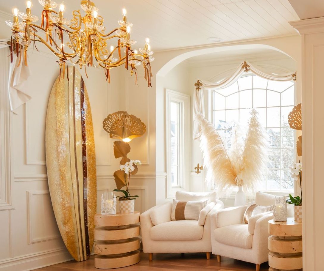 Luxurious living room with golden accents, surfboard, chandelier, and cream-colored furniture.