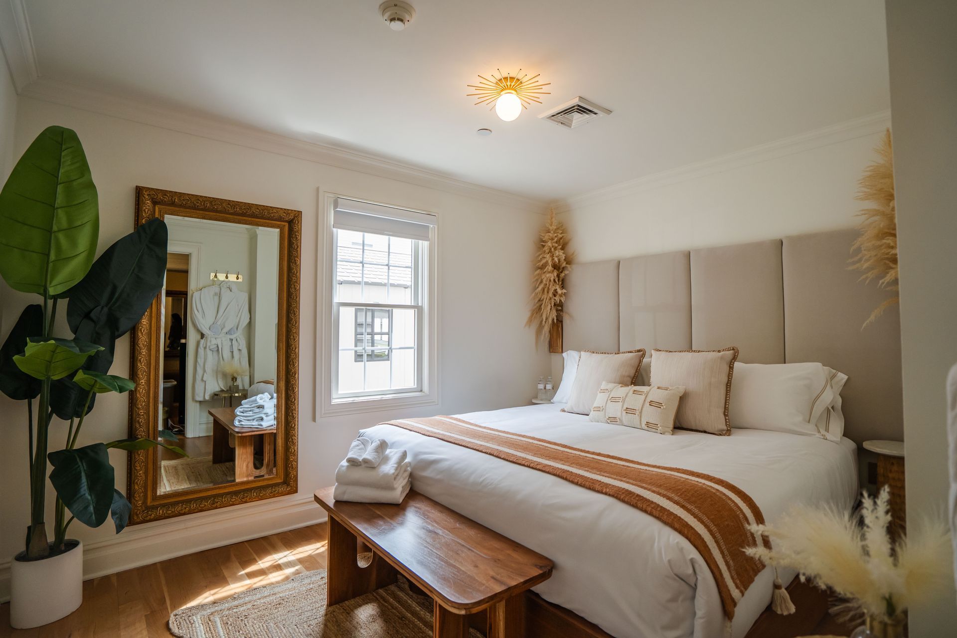 Bedroom with king-sized bed, large mirror, wooden bench, neutral tones, and plants.