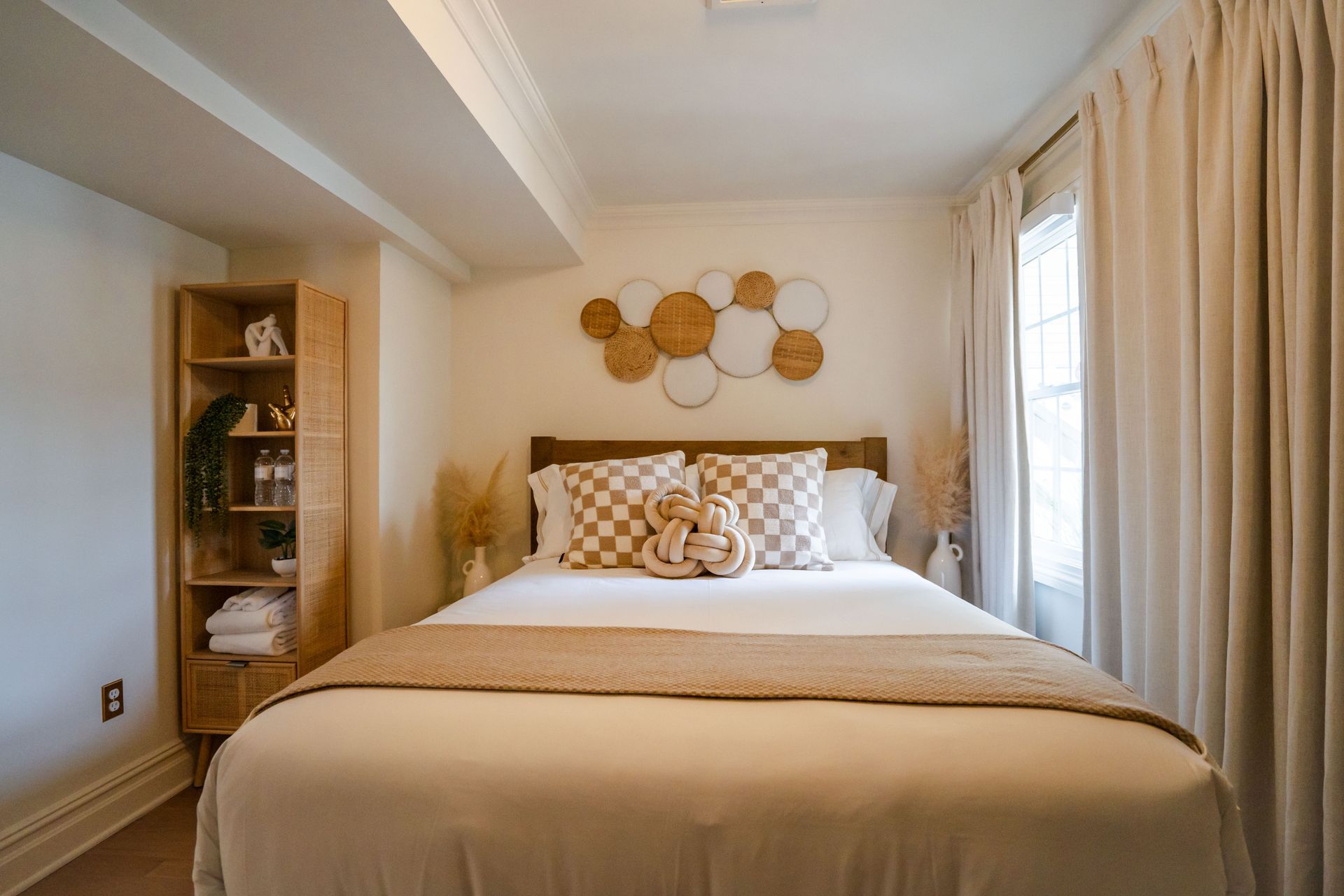 Bedroom with a beige bed, decorative pillows, wall decor, and a wooden cabinet. Beige curtains frame the window.