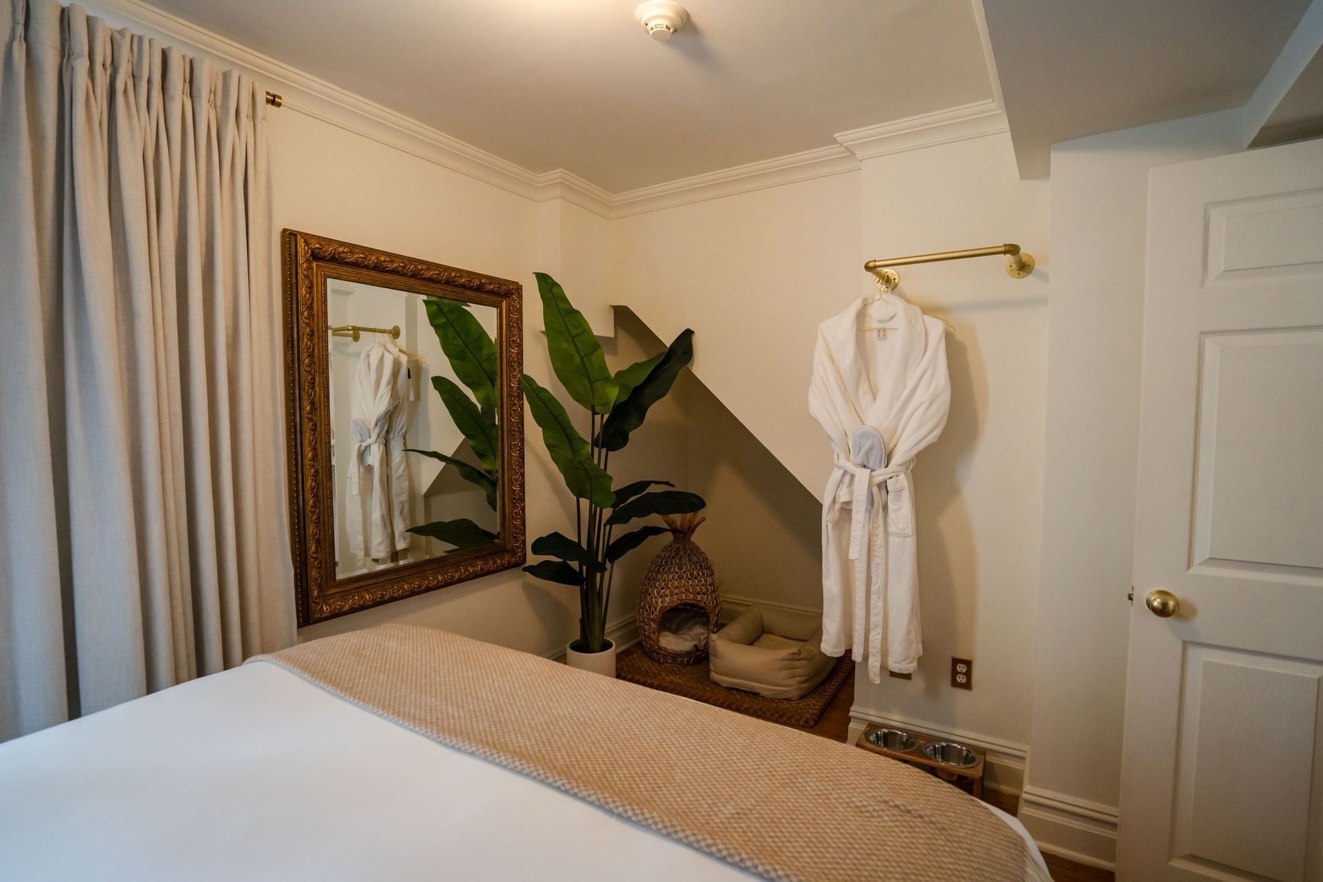 Bedroom with bed, mirror, robe, and plant against a white wall with beige curtains.
