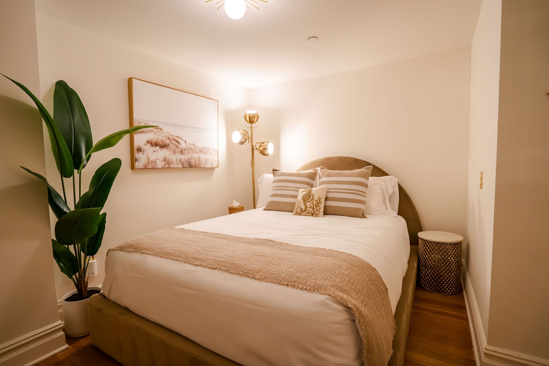 Cozy bedroom with bed, artwork, gold lamp, and tall plant. Beige and gold color scheme.