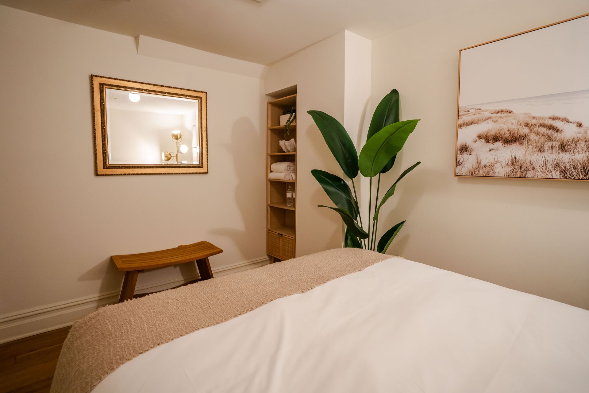 Massage room with a bed, bench, artwork, mirror, and plant.
