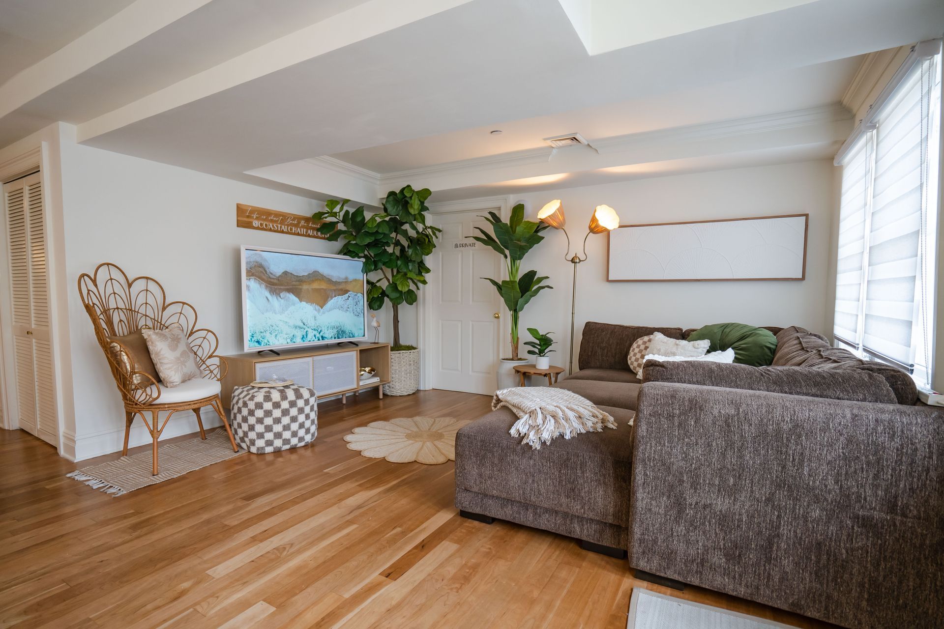 Living room with a brown sectional, wooden floor, large TV, and plants.