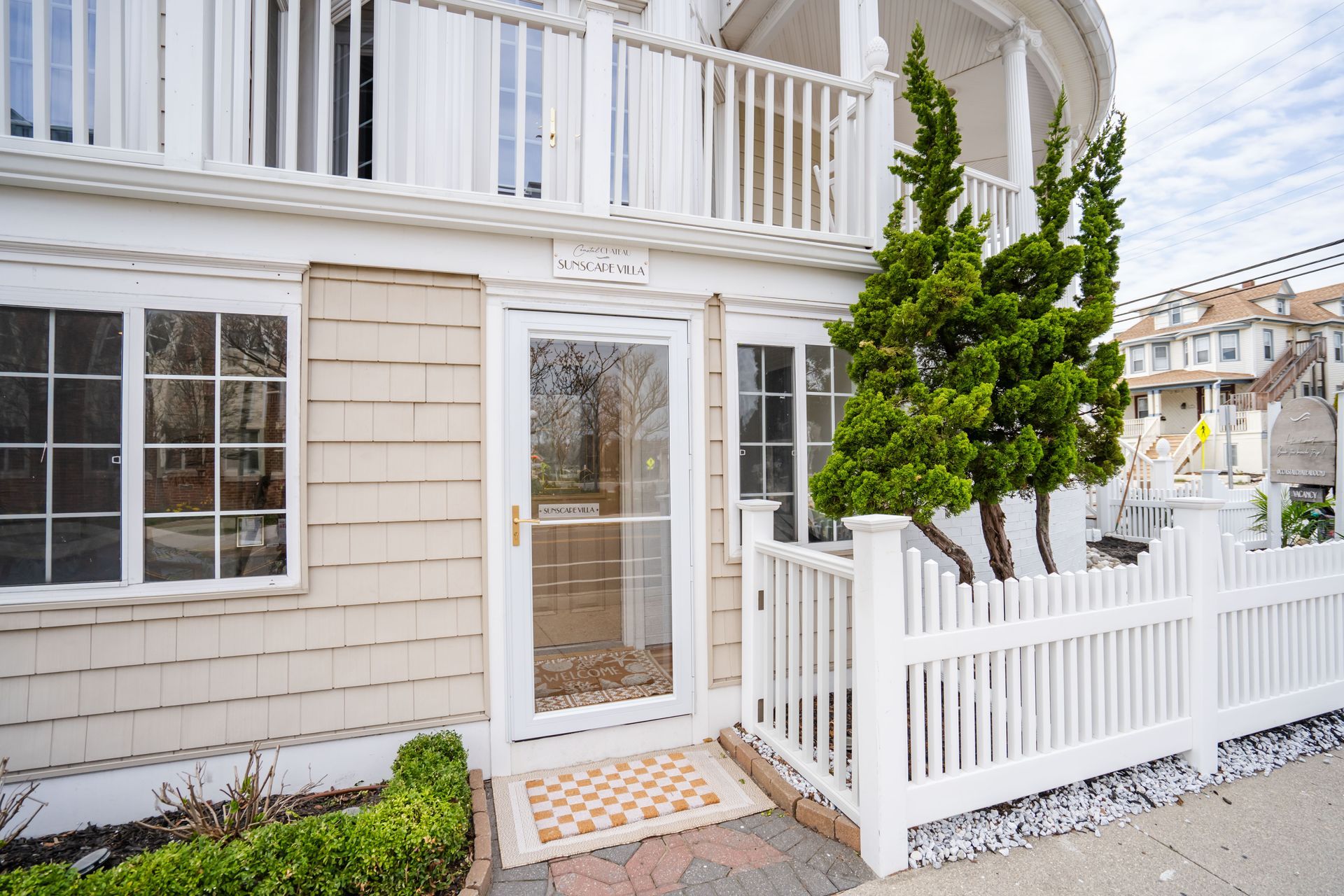 White building with picket fence, glass door, windows. A small evergreen tree grows nearby.