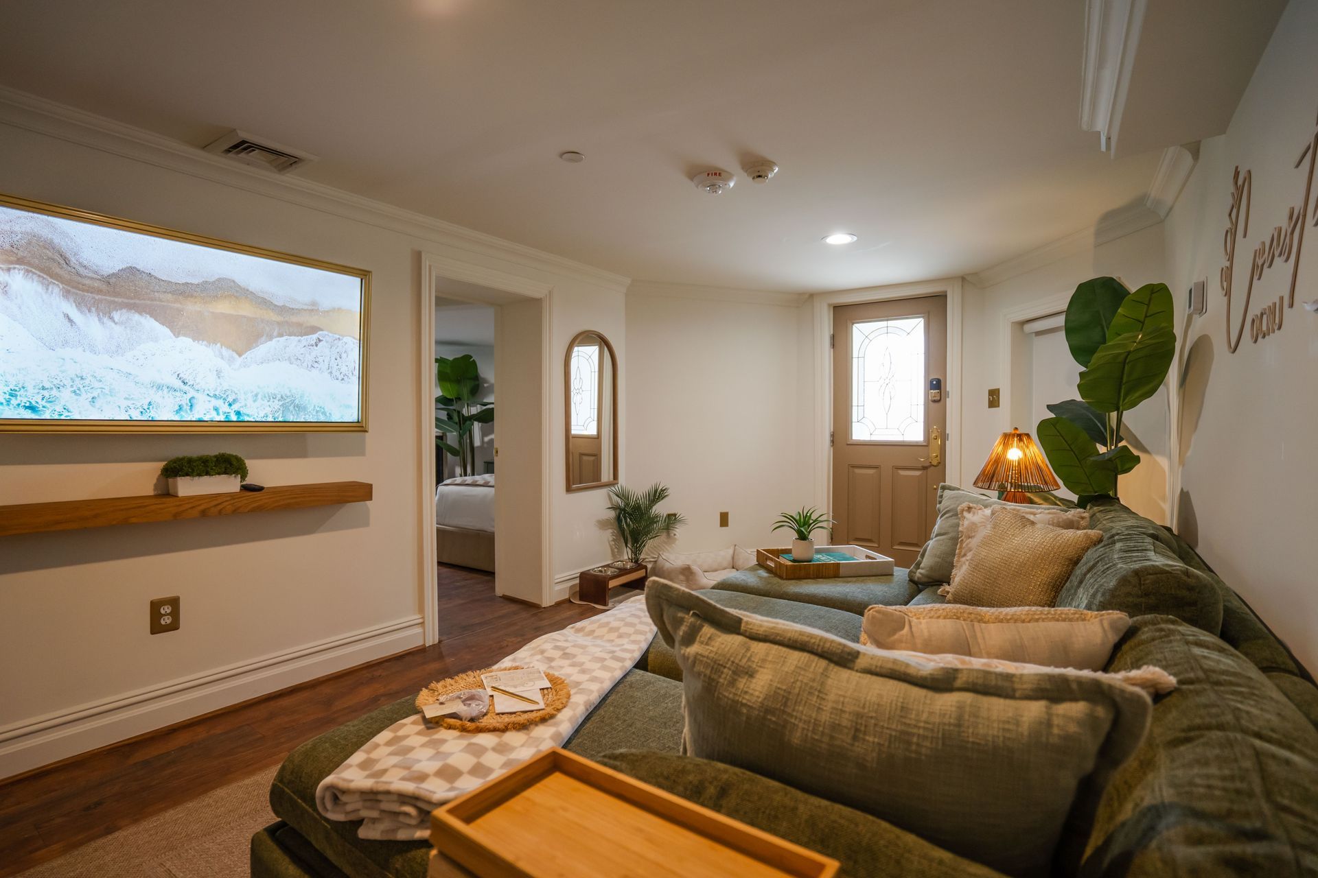 Cozy living room with green sofa, wooden accents, and a mounted TV. Doorway leads to a bedroom.