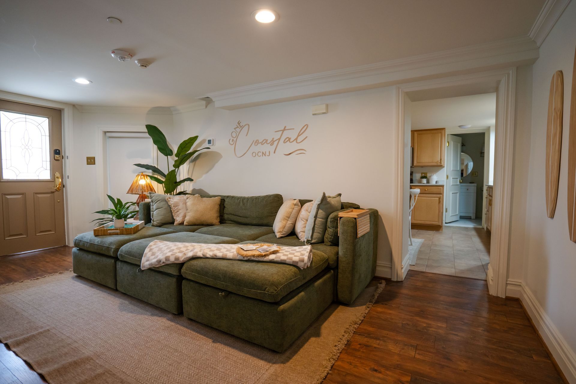 Green sectional sofa in a living room, rug, wooden floor, open doorway to a kitchen.