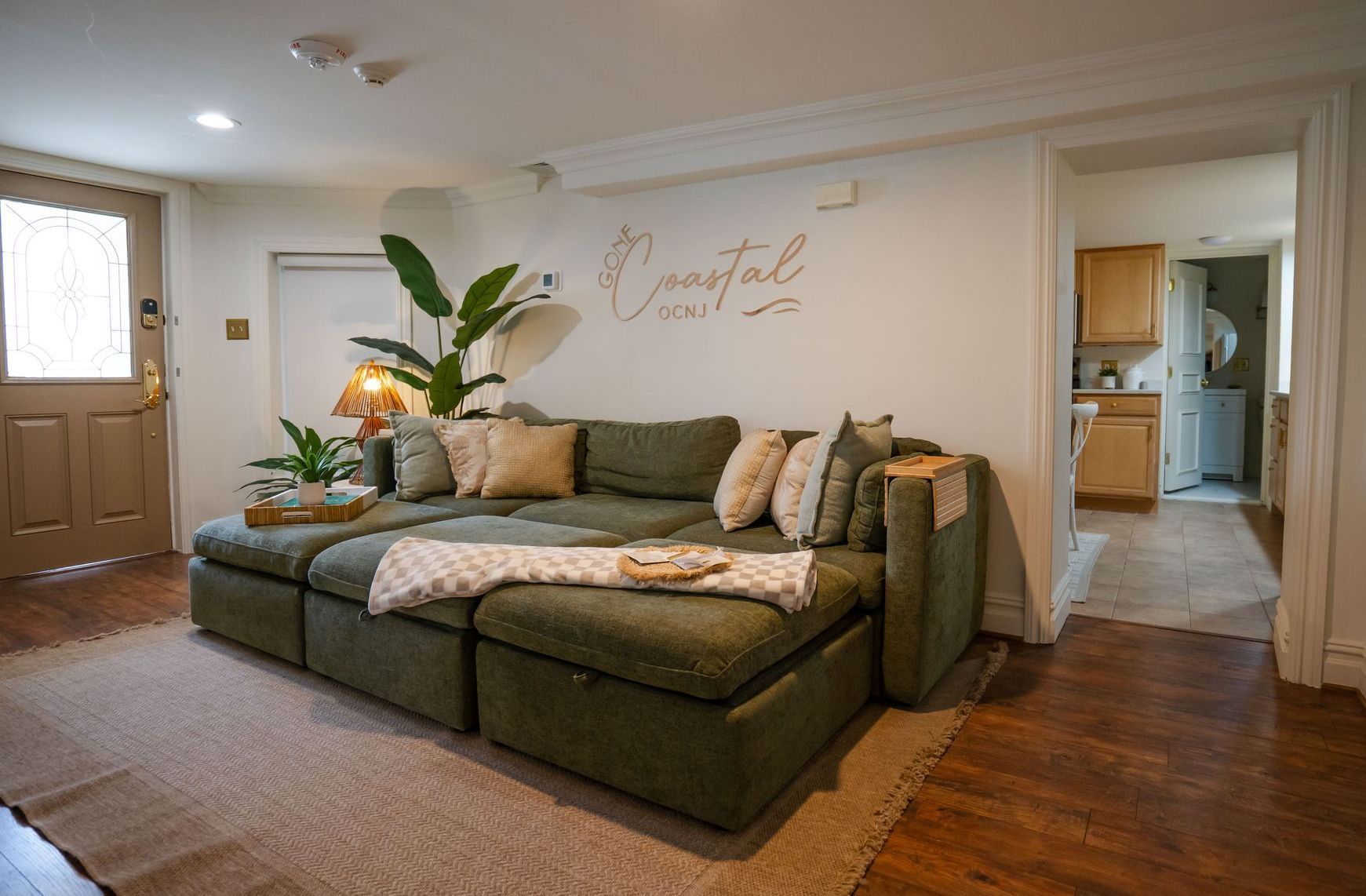Green sectional sofa in a living room with plants, a door, and a glimpse into a kitchen.