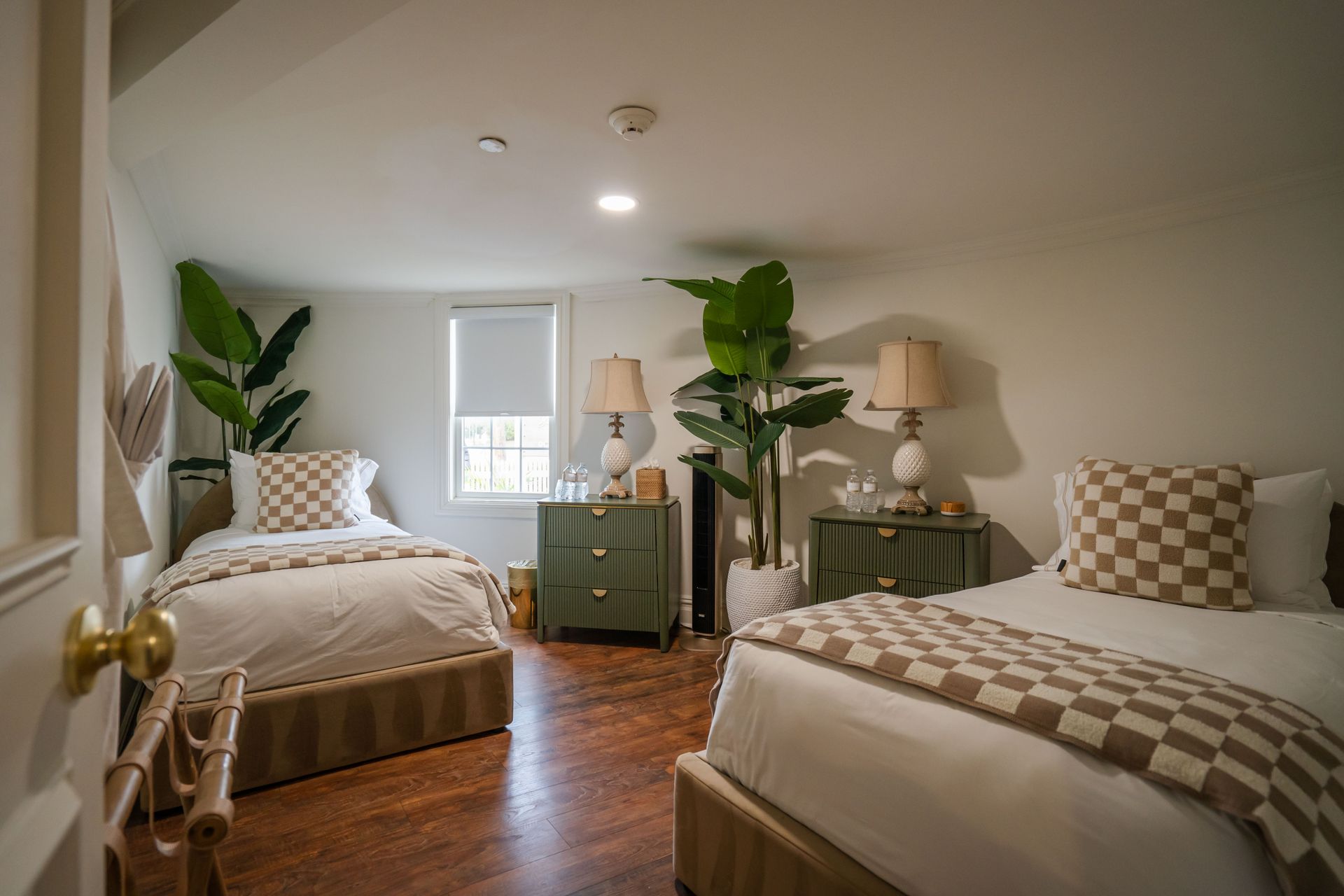 Bedroom with two beds, olive green bedside tables, plants, and checkered throw blankets.