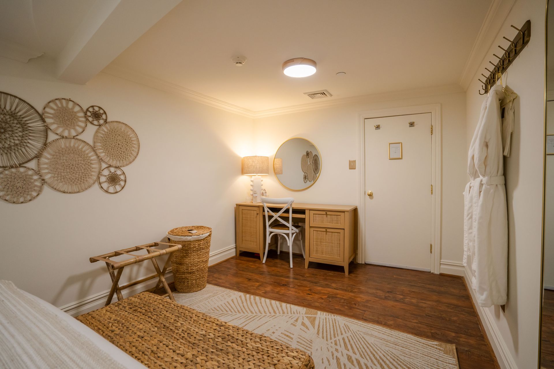 Cozy bedroom with wicker desk, mirror, and wall decor; a door, robes, and a rug on wood floor.