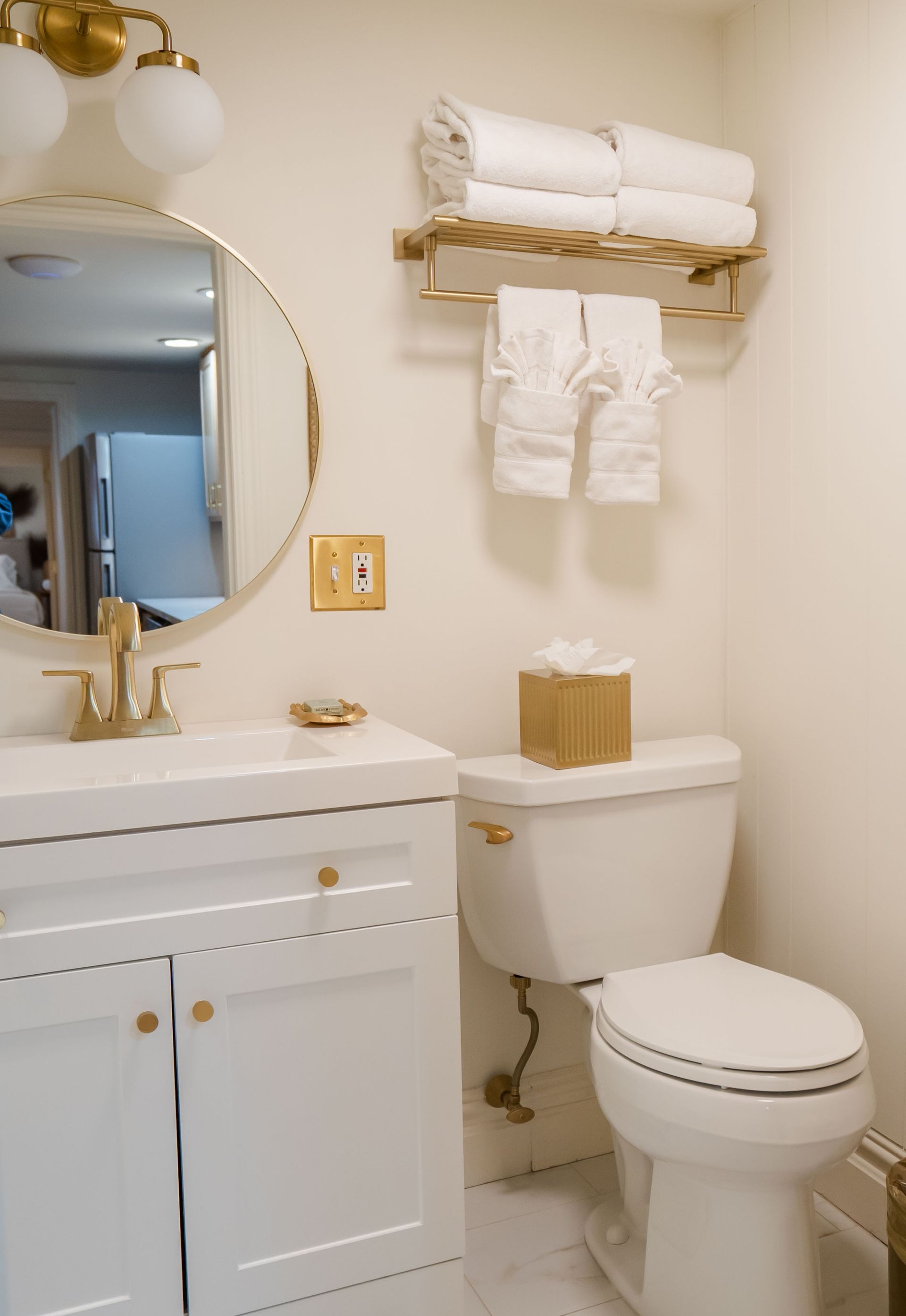 White bathroom with gold accents: vanity, mirror, toilet, towel rack, and tissue box.