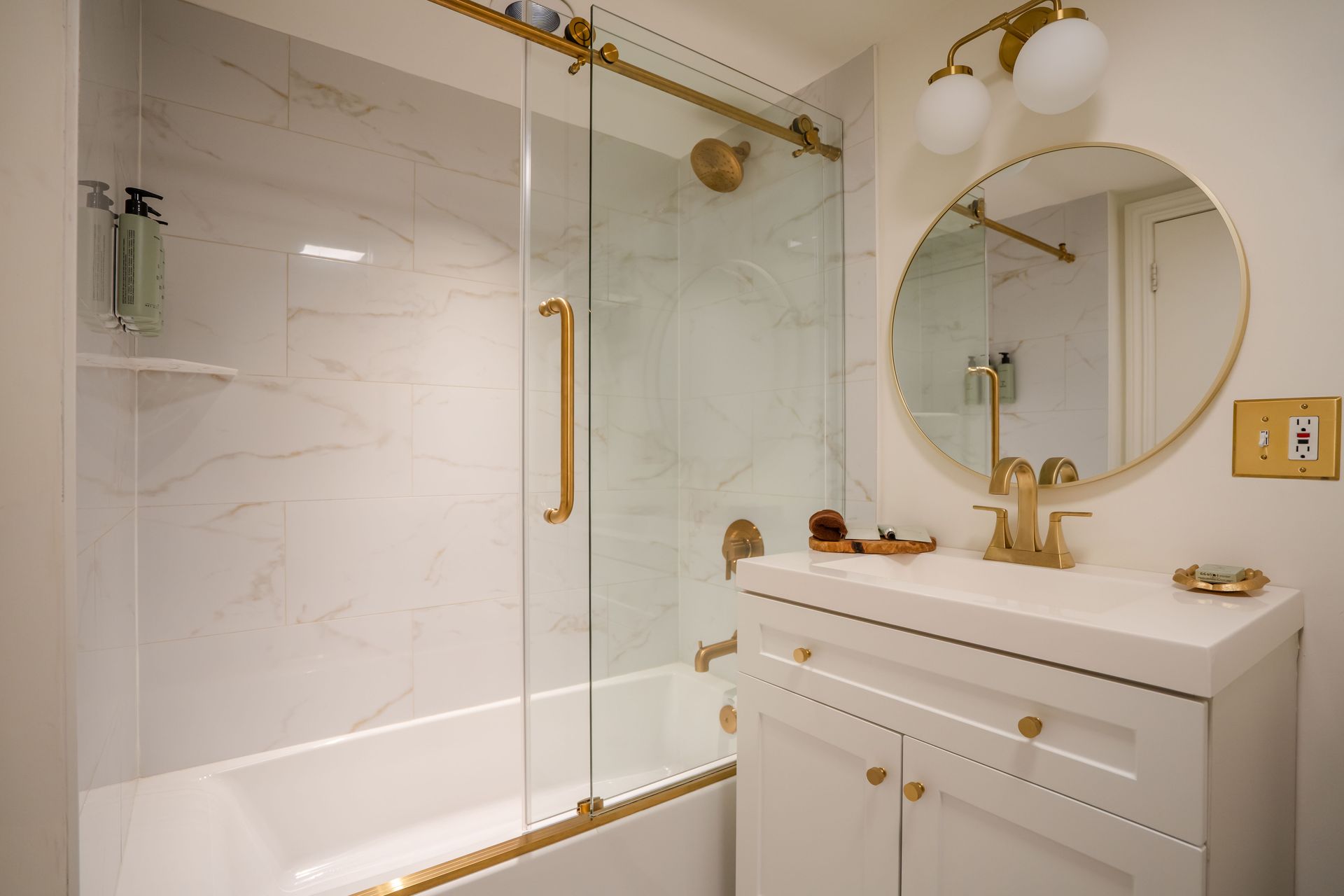 Bathroom with white and gold accents, including a tub, shower, vanity, and round mirror.
