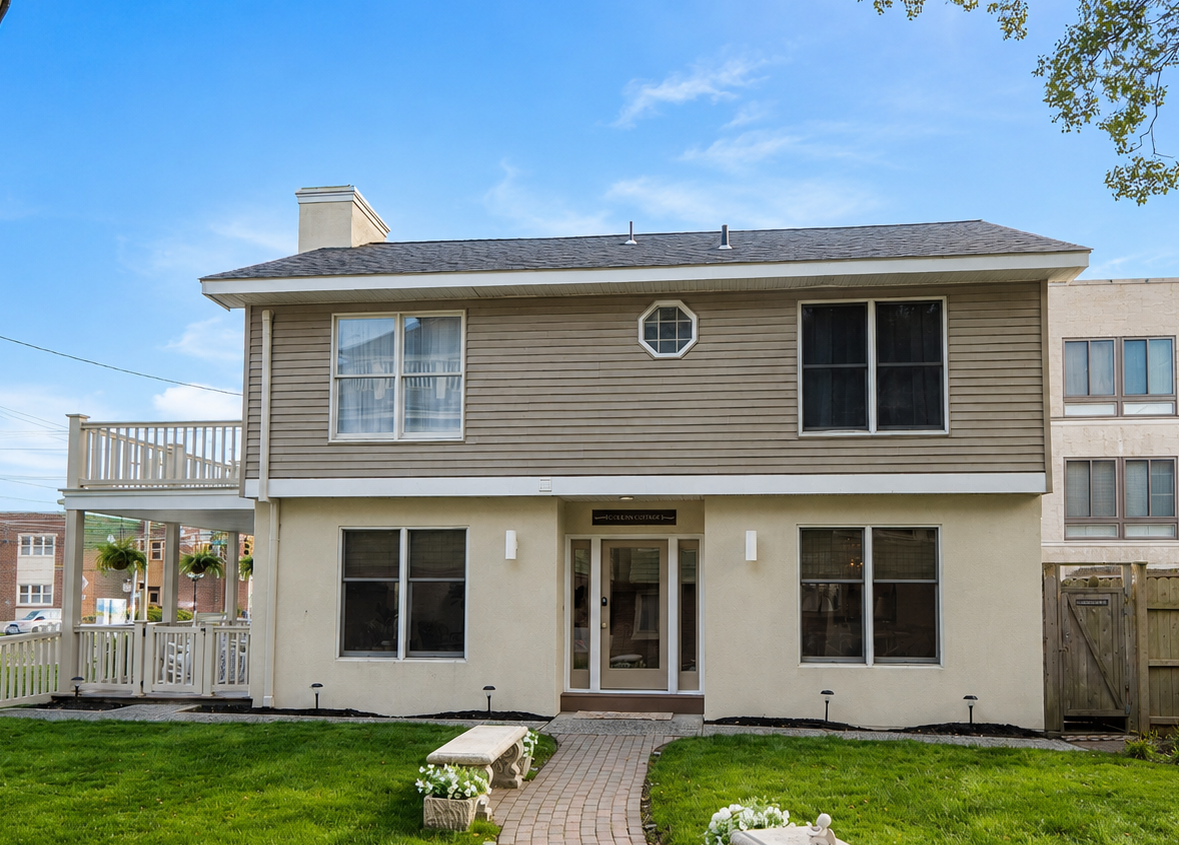 Two-story beige house with a small balcony, front lawn, and stone walkway under a blue sky