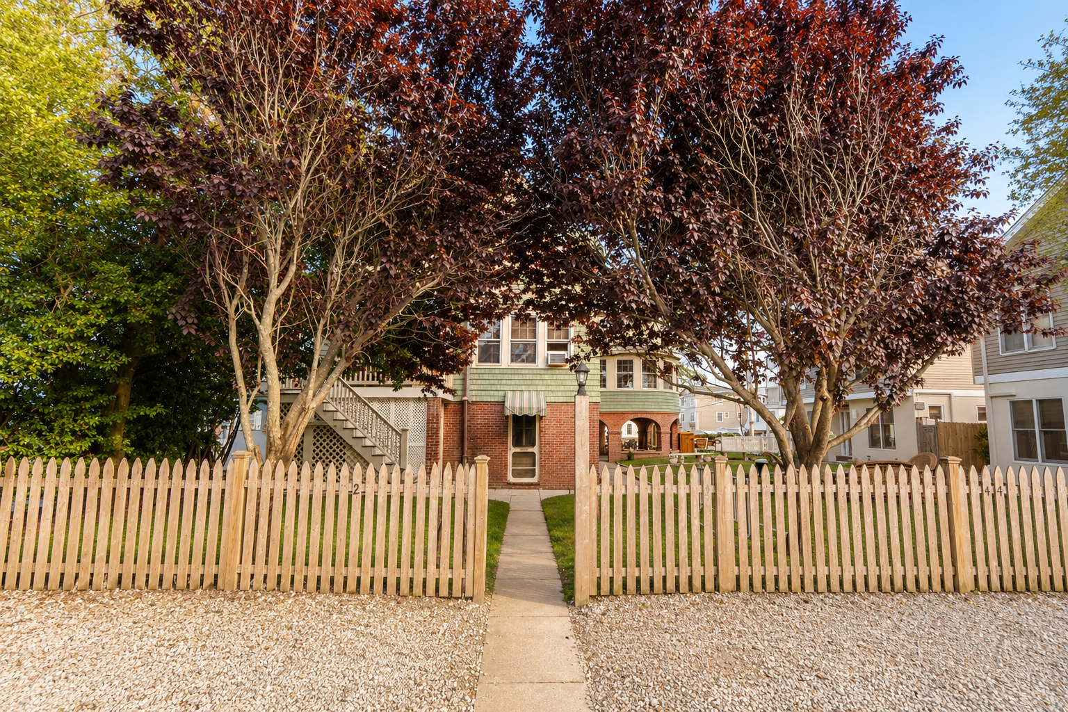 Brick house behind a tan picket fence, framed by trees and a gravel front yard.