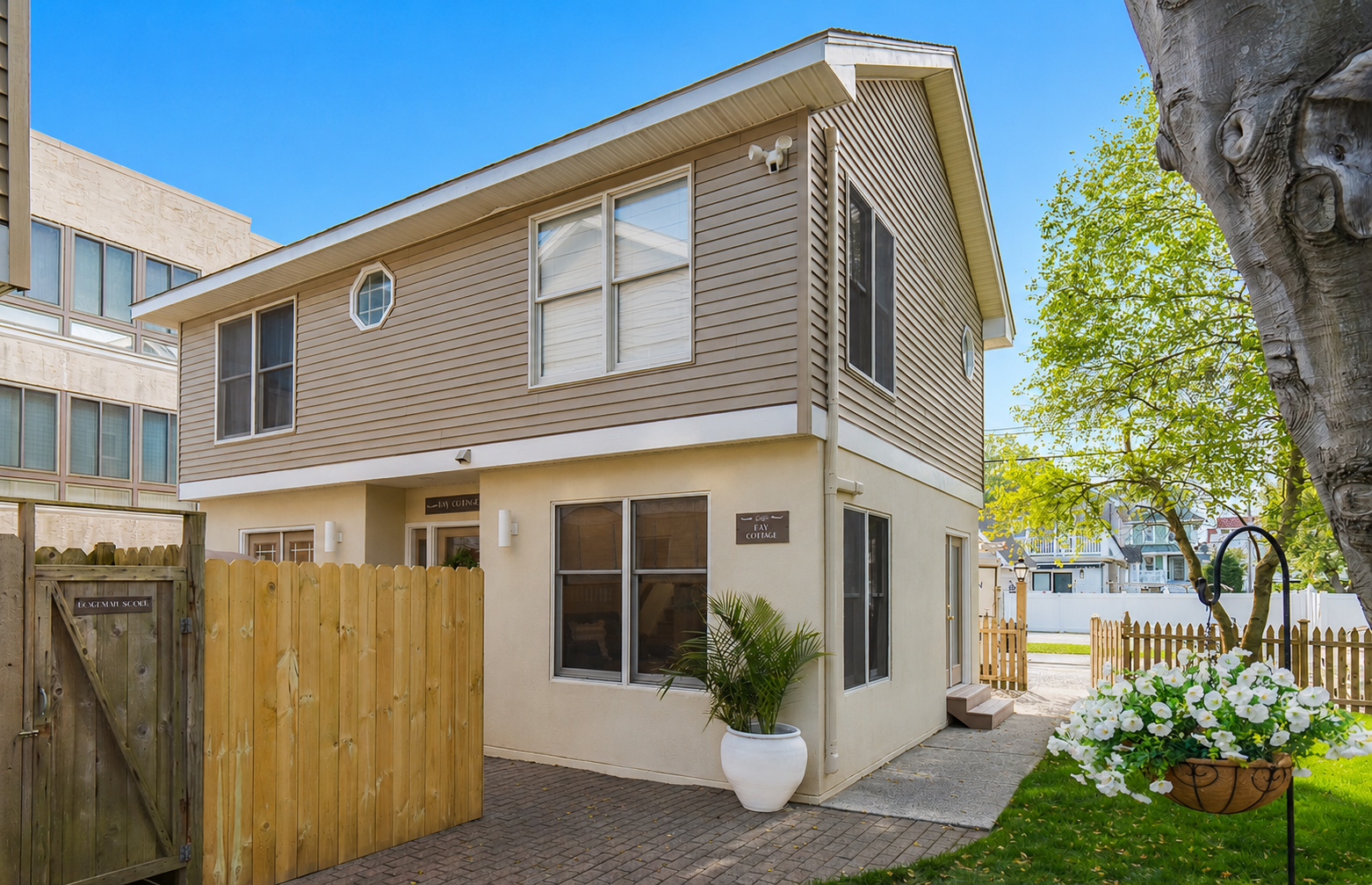 Two-story beige house with wood fence, potted plant, and green yard in a sunny residential area
