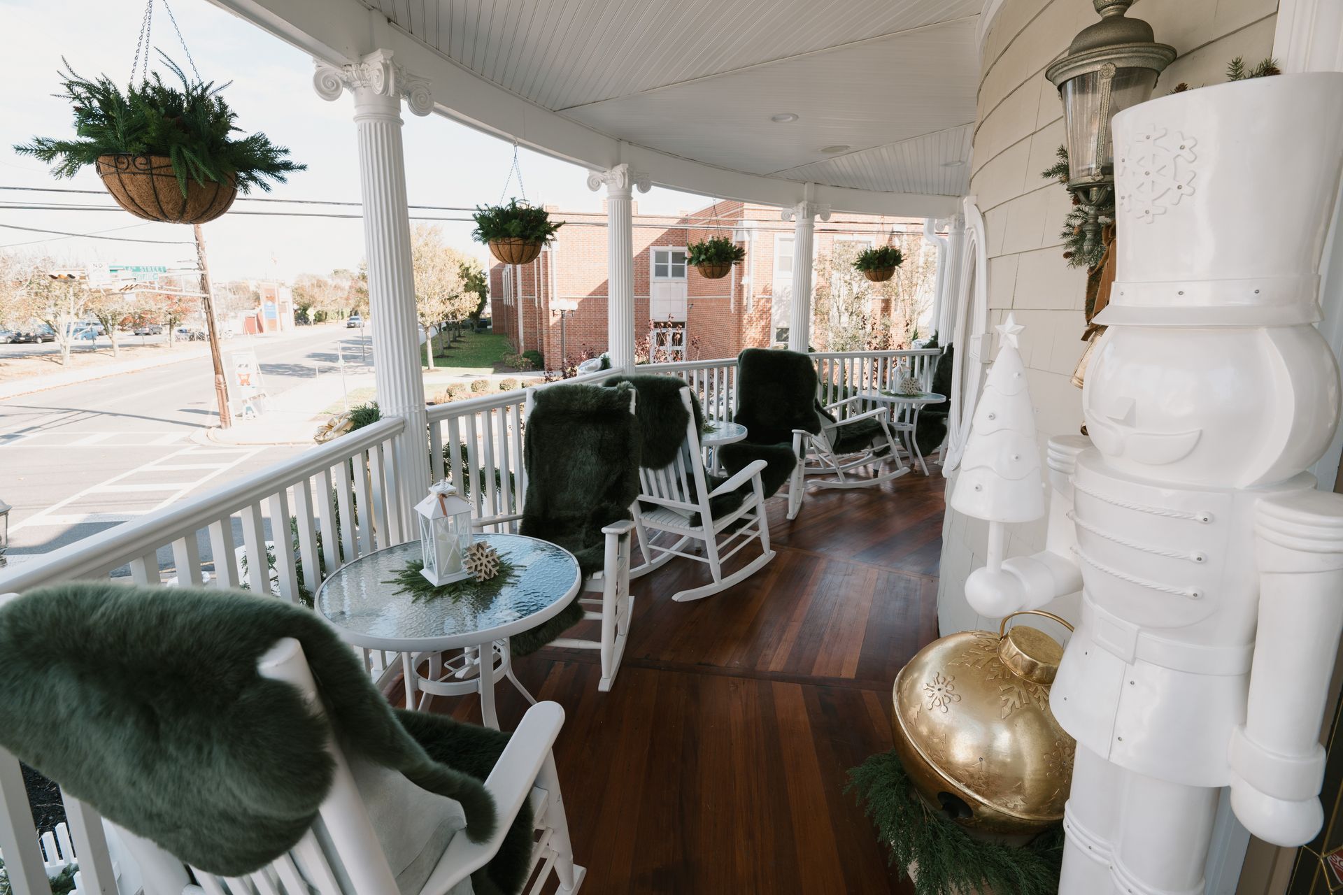 White porch with rocking chairs and Christmas decorations, overlooking a street and building.