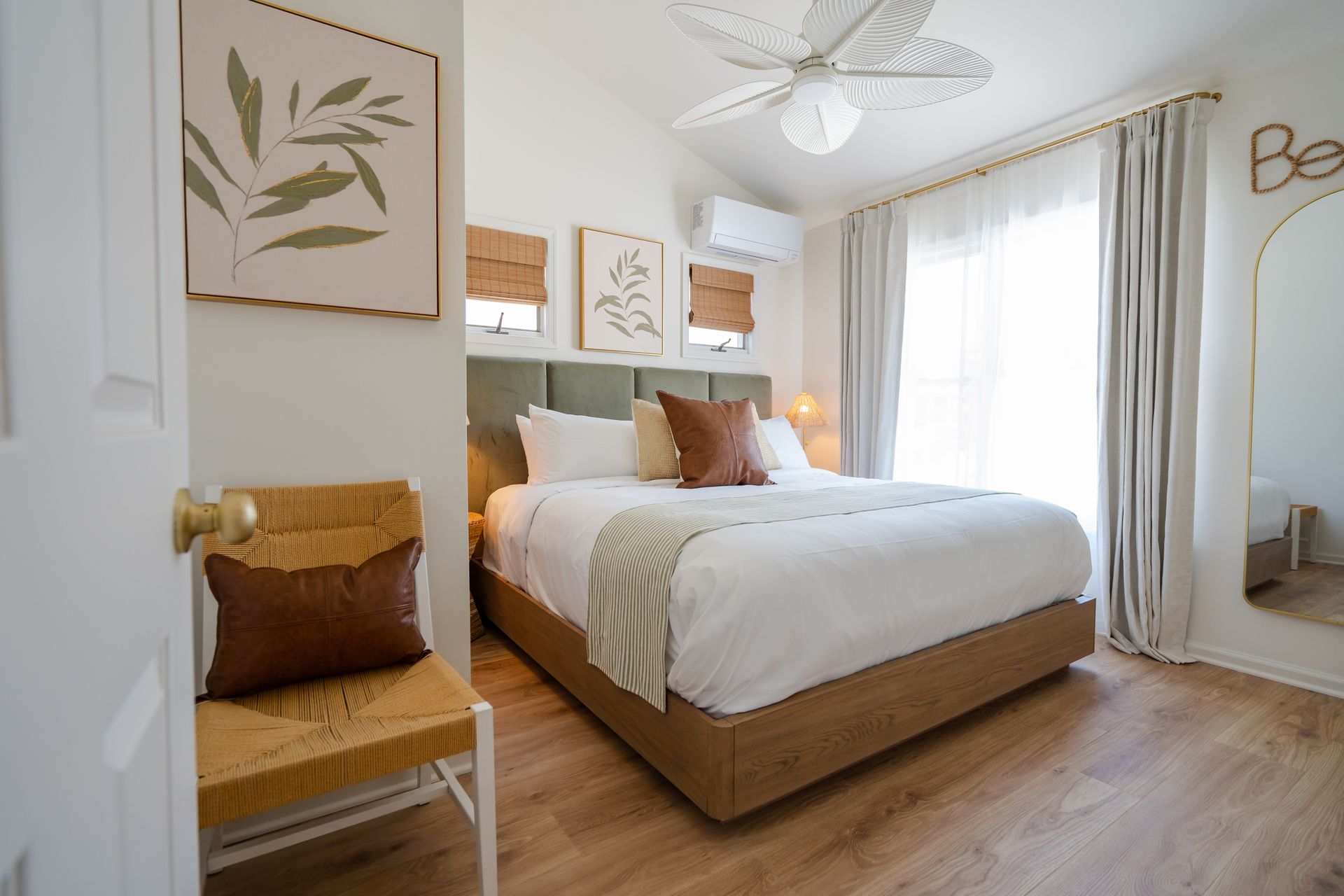 Bright, airy bedroom with a bed, wicker chair, wall art, and a large sunlit window