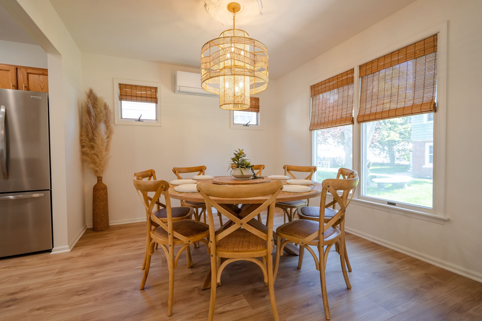 Bright dining room with a round wooden table, woven chairs, chandelier, and large windows with bamboo blinds