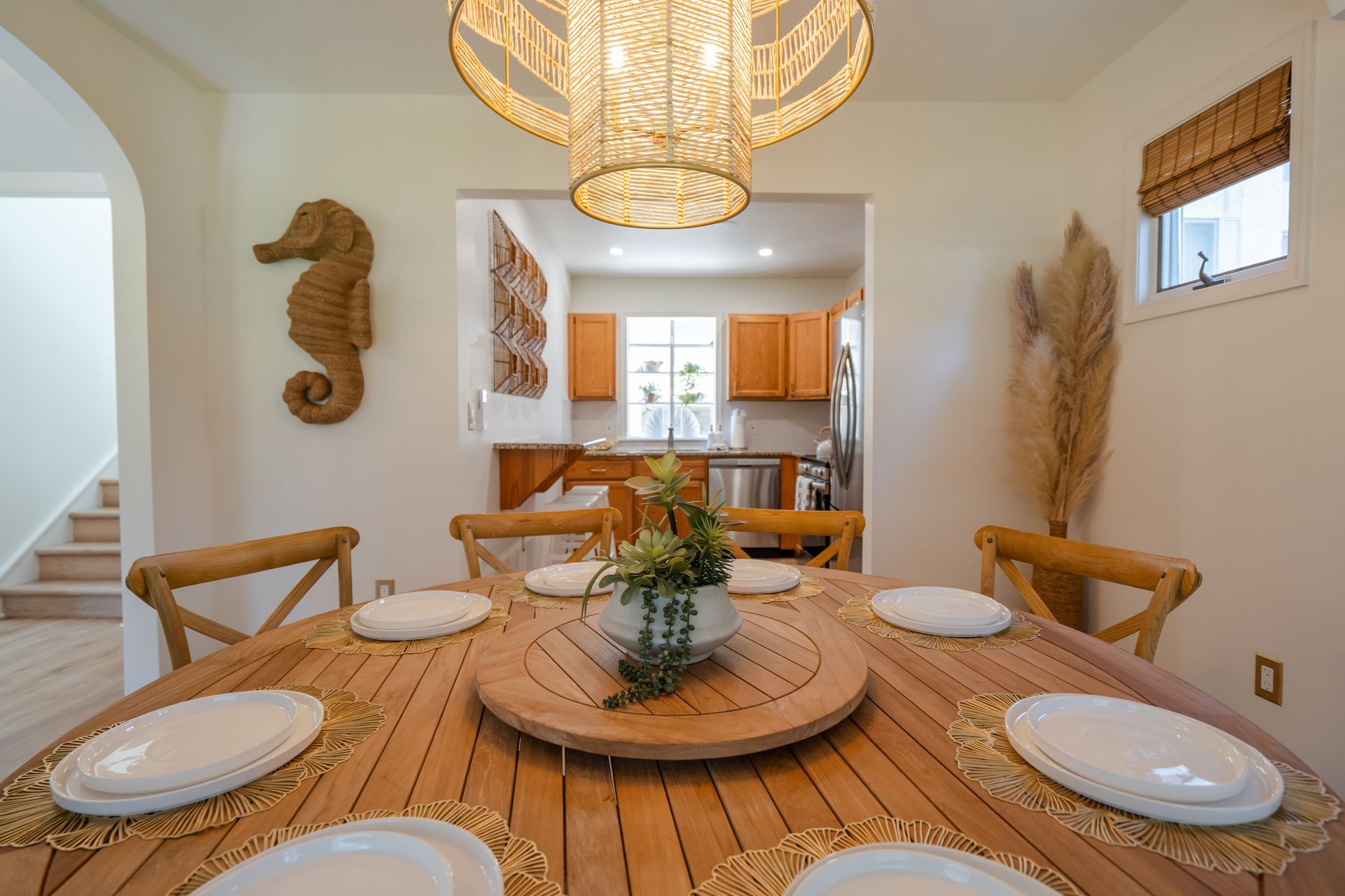 Bright dining room with a round wooden table, woven chairs, hanging pendant light, and a view into the kitchen