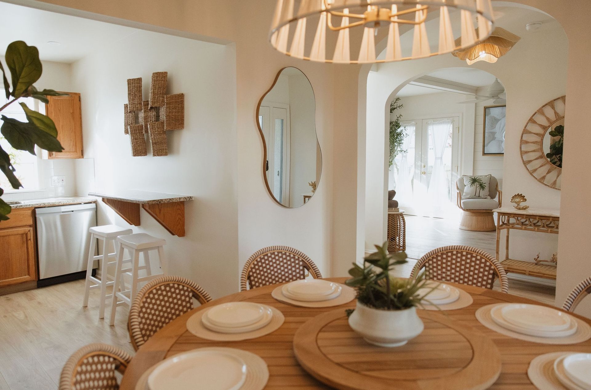 Bright dining room with round wooden table, woven chairs, neutral decor, and a kitchen opening on the left