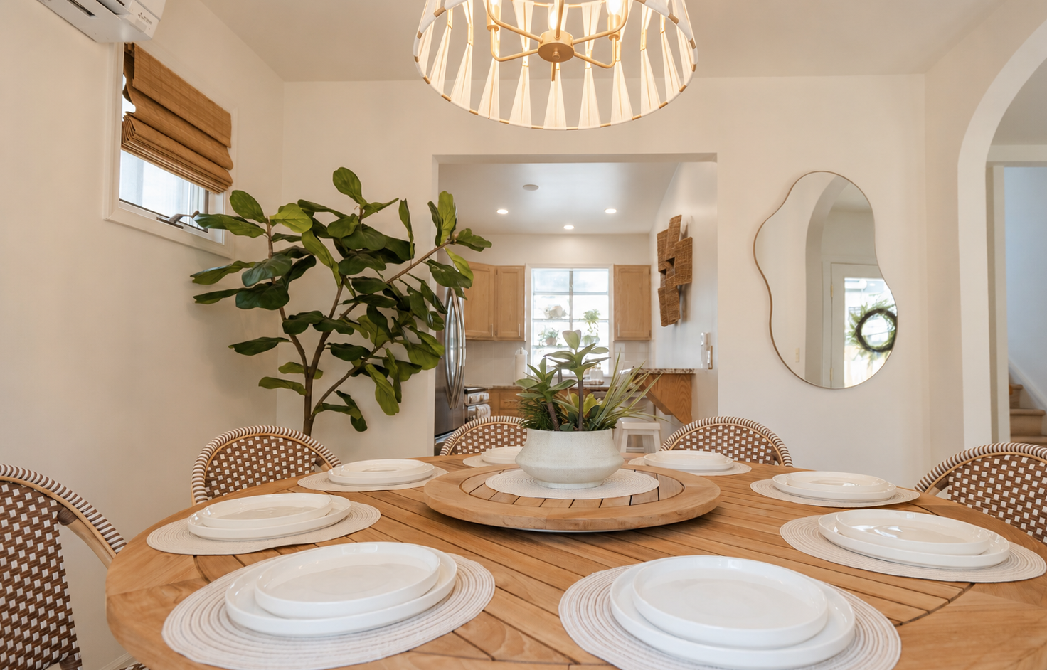 Bright dining room with a round table, white plates, woven chairs, and a large plant