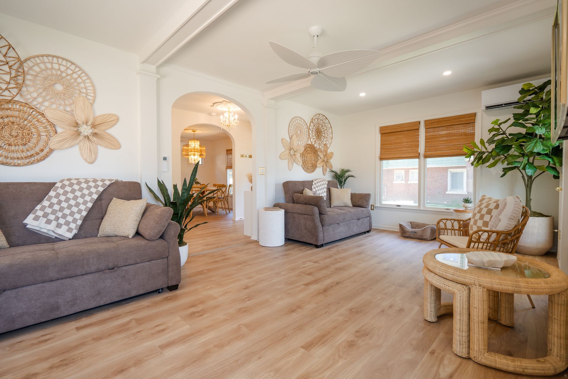 Bright living room with gray sofas, wood floors, wicker chair, plants, and wall art under white ceilings