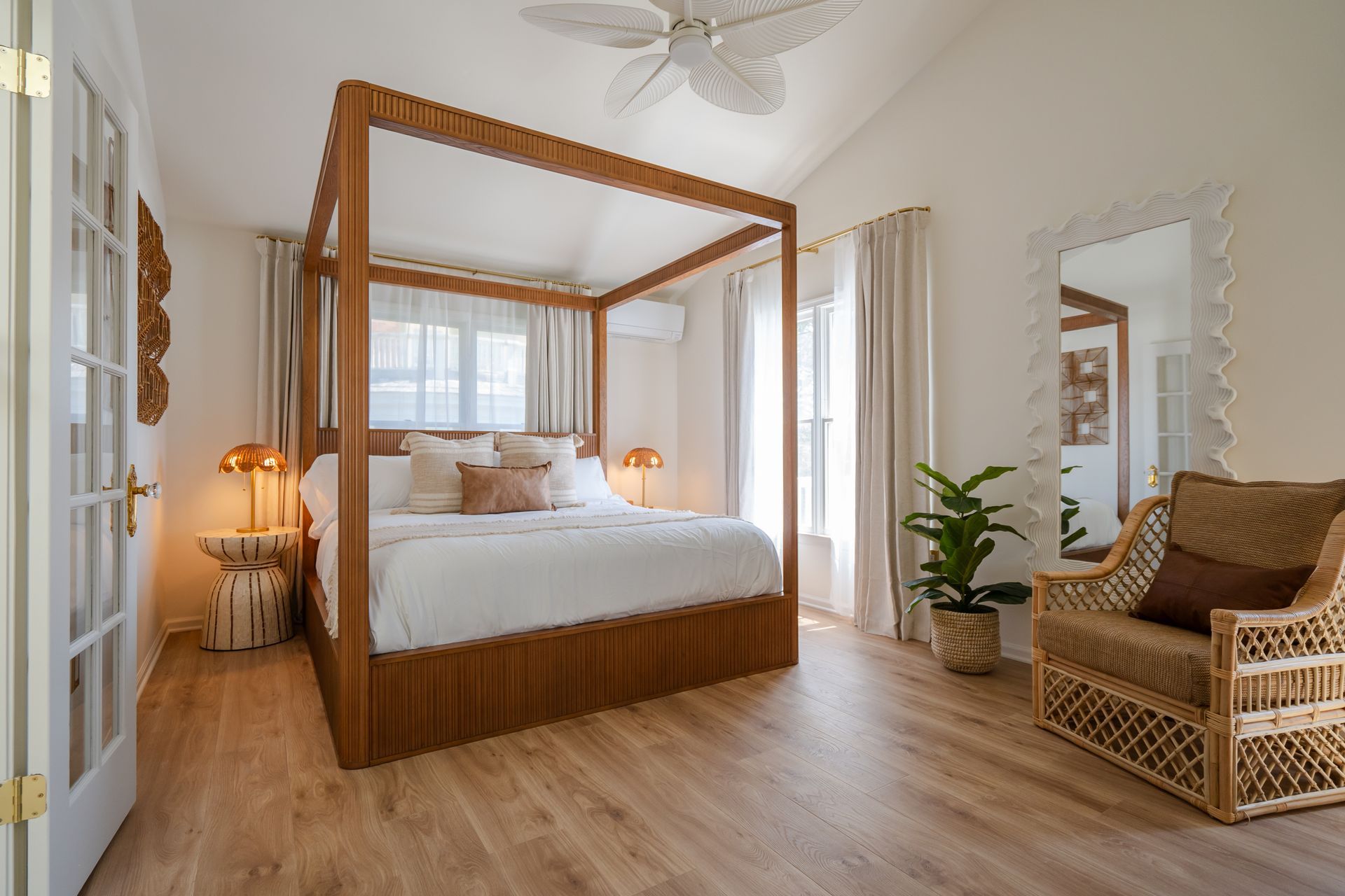 Bright bedroom with a wooden canopy bed, wicker chair, lamps, plants, and large windows.