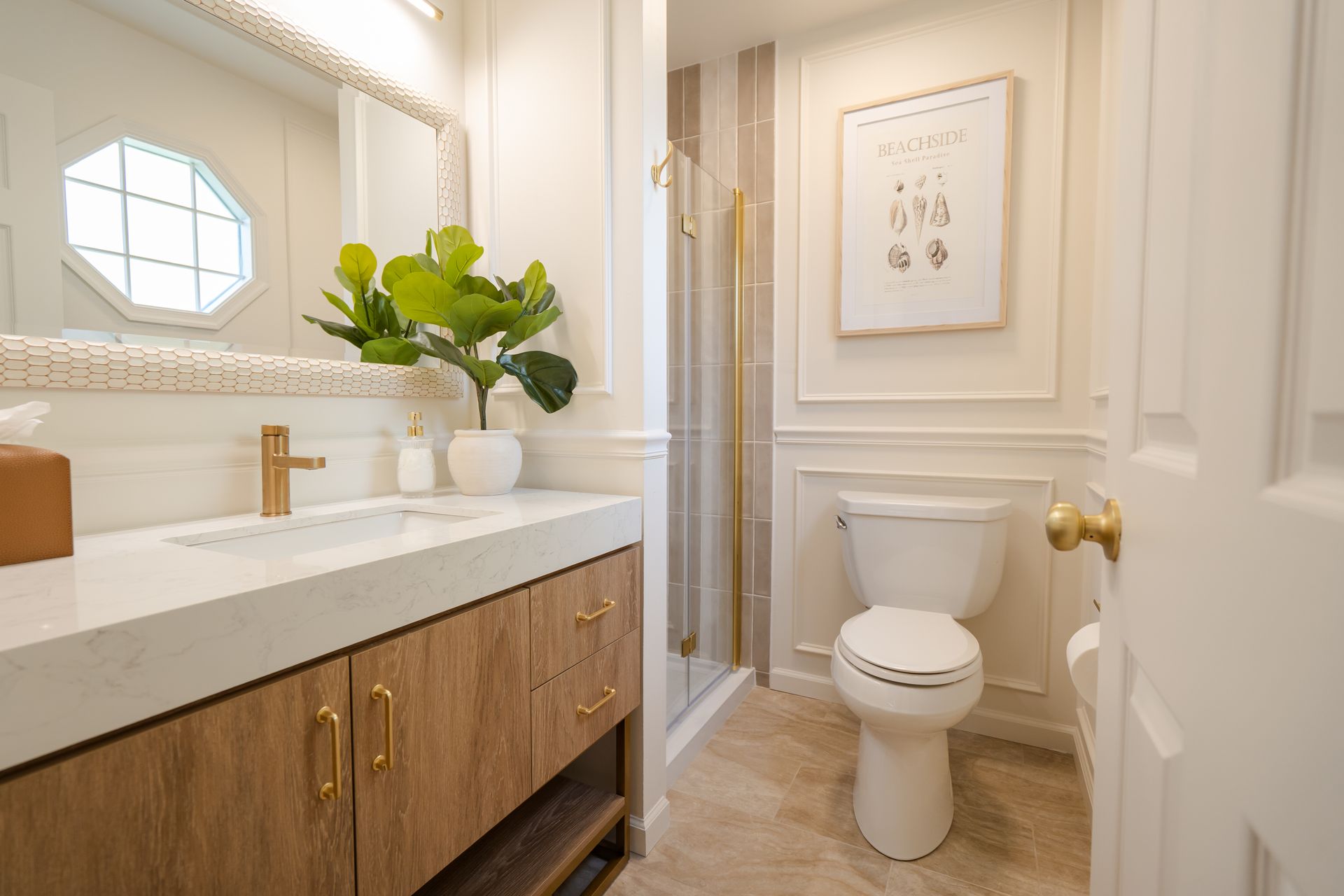 Bright bathroom with vanity, brass fixtures, round mirror, potted plant, and a toilet beside a glass shower.