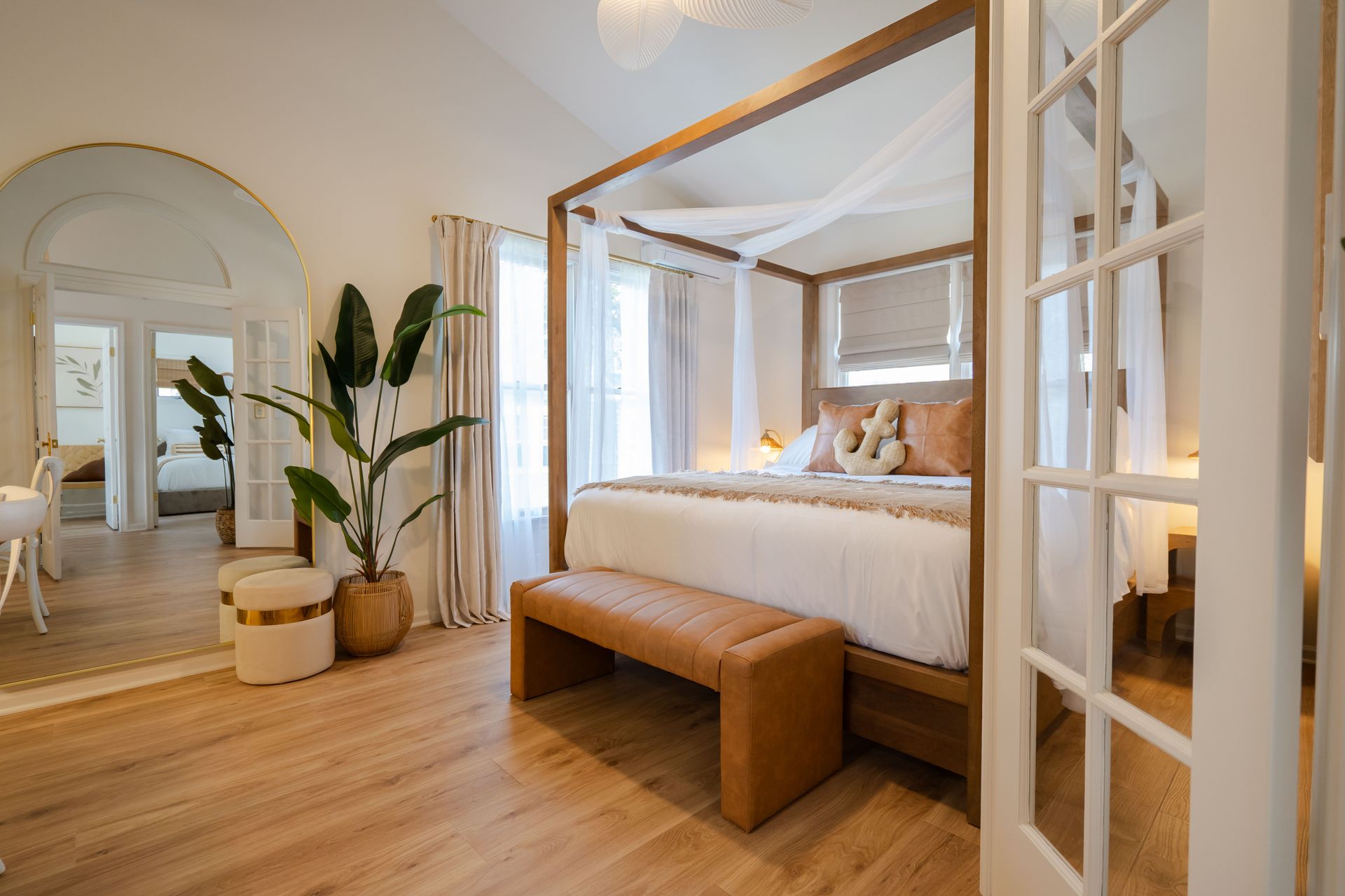 Bright bedroom with a four-poster bed, wooden bench, large mirror, and potted plant near French doors.