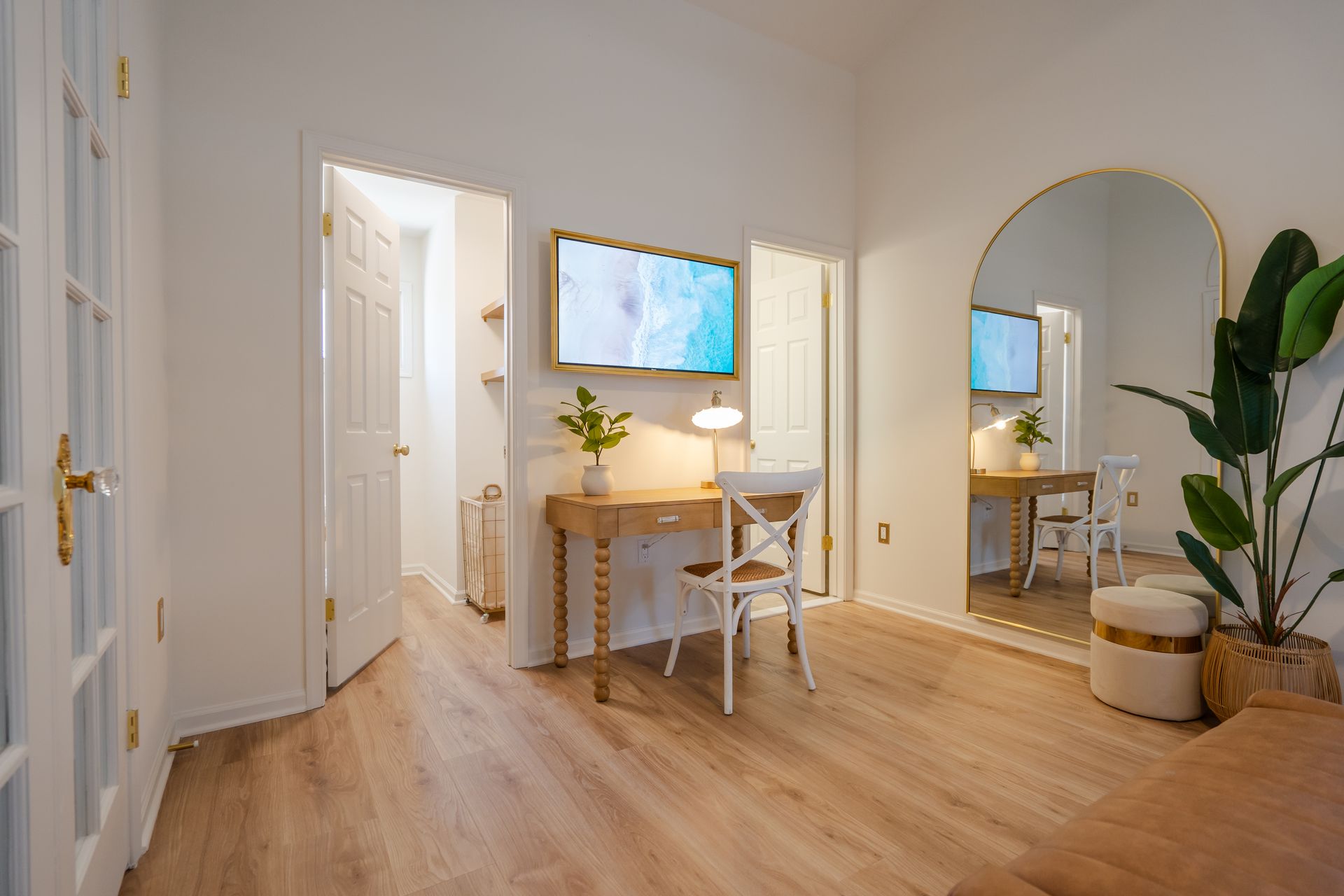 Bright hallway office with desk, chair, wall art, round mirror, and a large plant on wood floors.