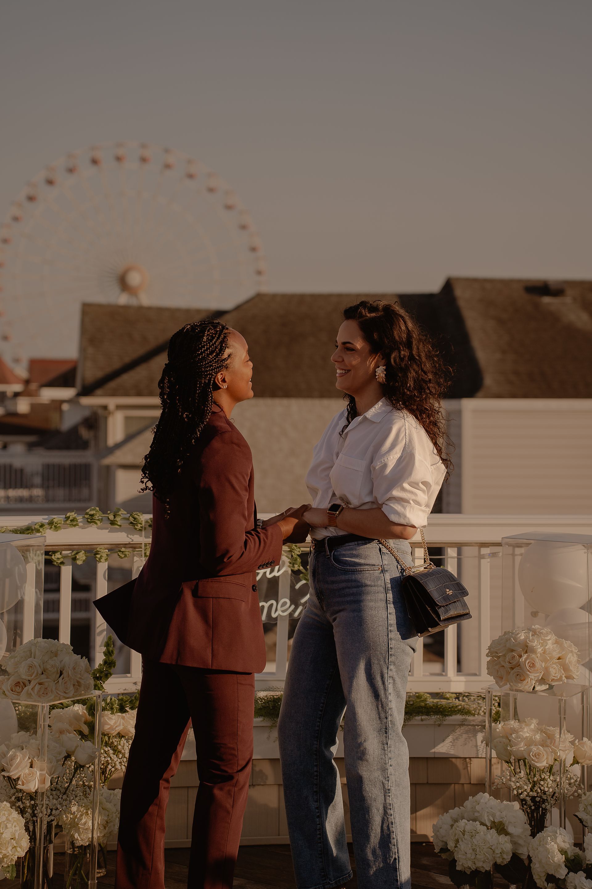 Two women holding hands, facing each other on a rooftop. White flowers and a Ferris wheel in the background.