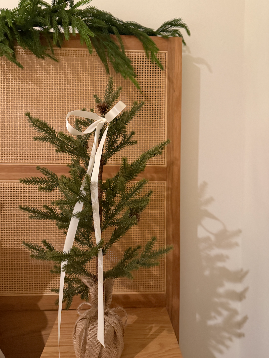 Small Christmas tree with white ribbon, burlap base. Set against a wooden backdrop with evergreen garland.