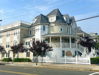 Victorian-style building with white picket fence, round porch, and dark roof. Trees and power lines in front.