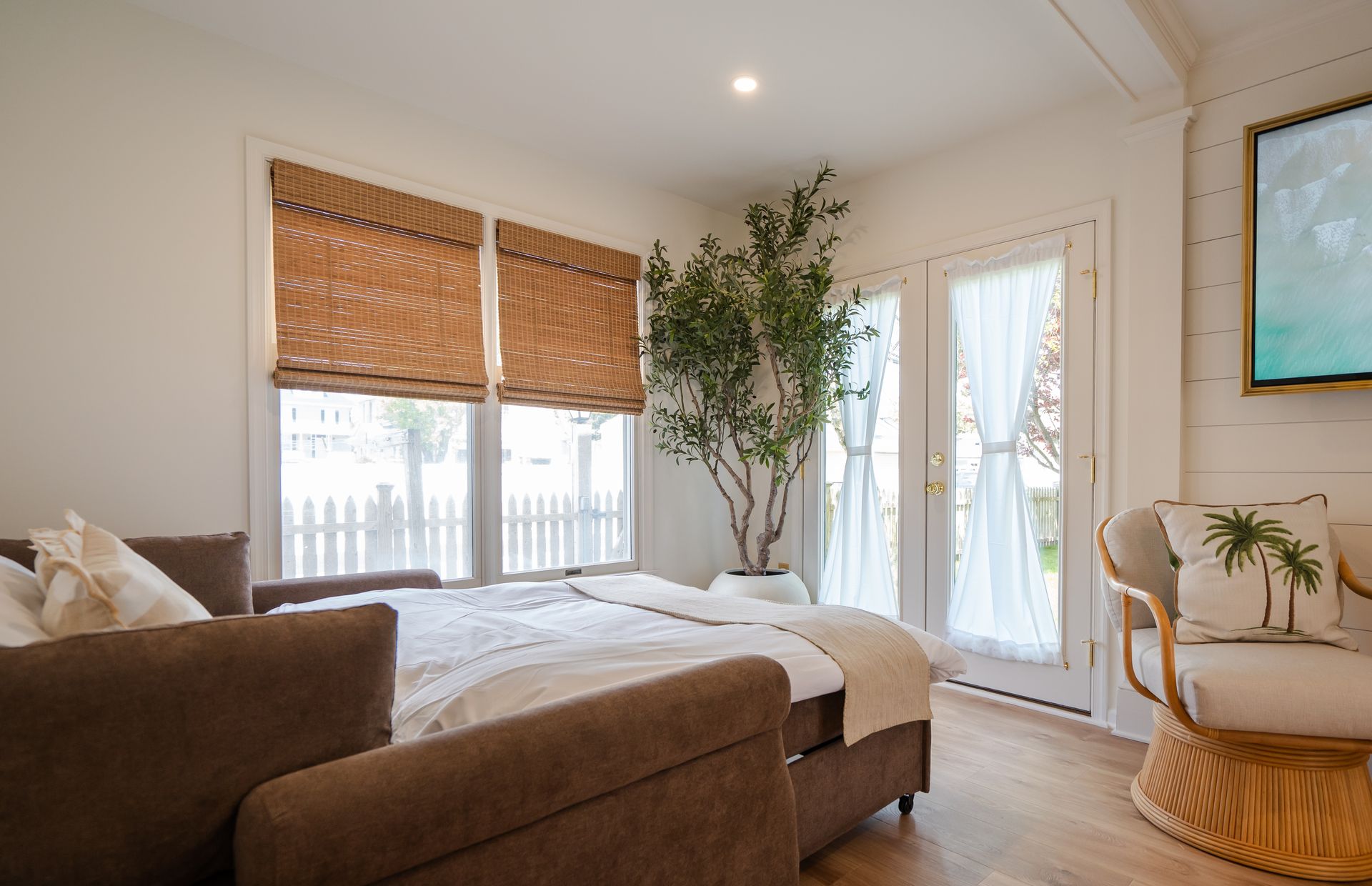 Bright living room with sofa, bed, large windows, potted tree, and a wicker chair near a glass door.