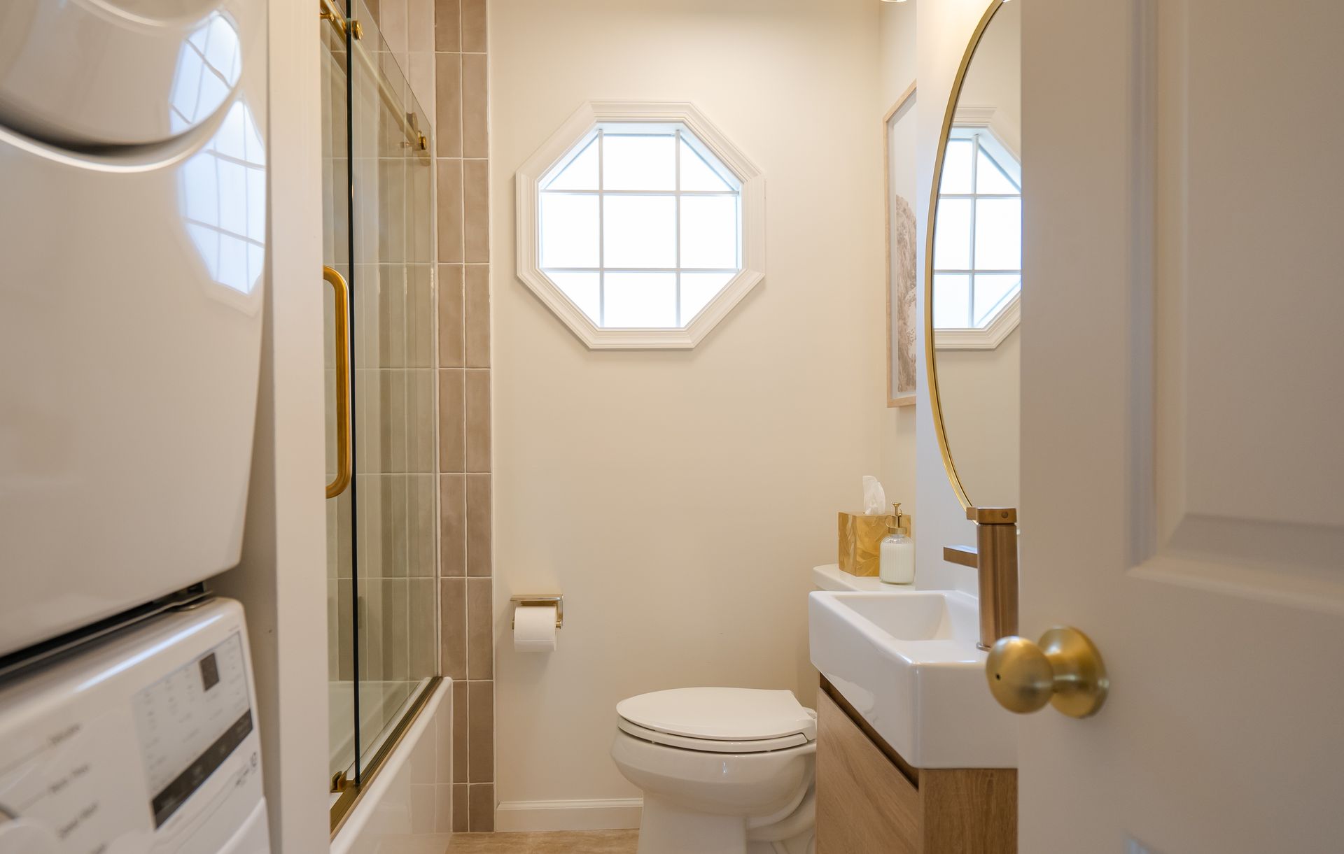 Compact bathroom with toilet, shower, sink, octagonal window, and stacked washer and dryer.