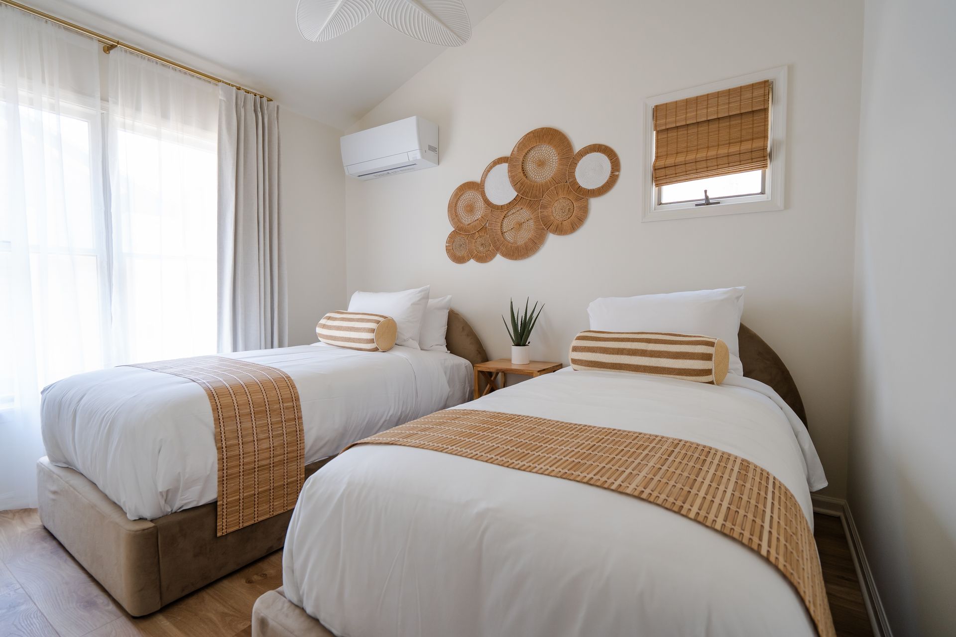 Bright bedroom with two single beds, woven wall decor, and a window with curtains and bamboo shade