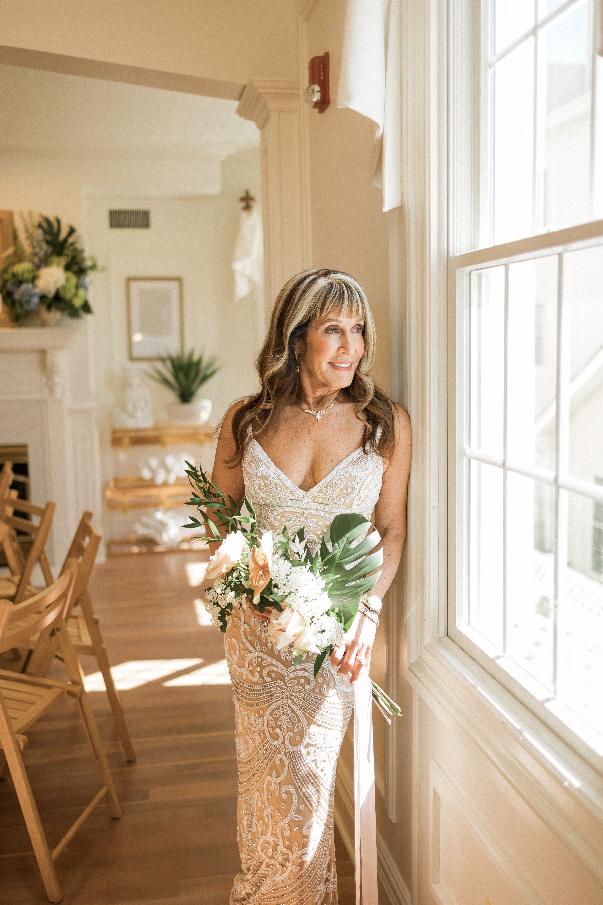 Woman in sequined wedding dress holding flowers, leaning against a sunlit window, smiling.