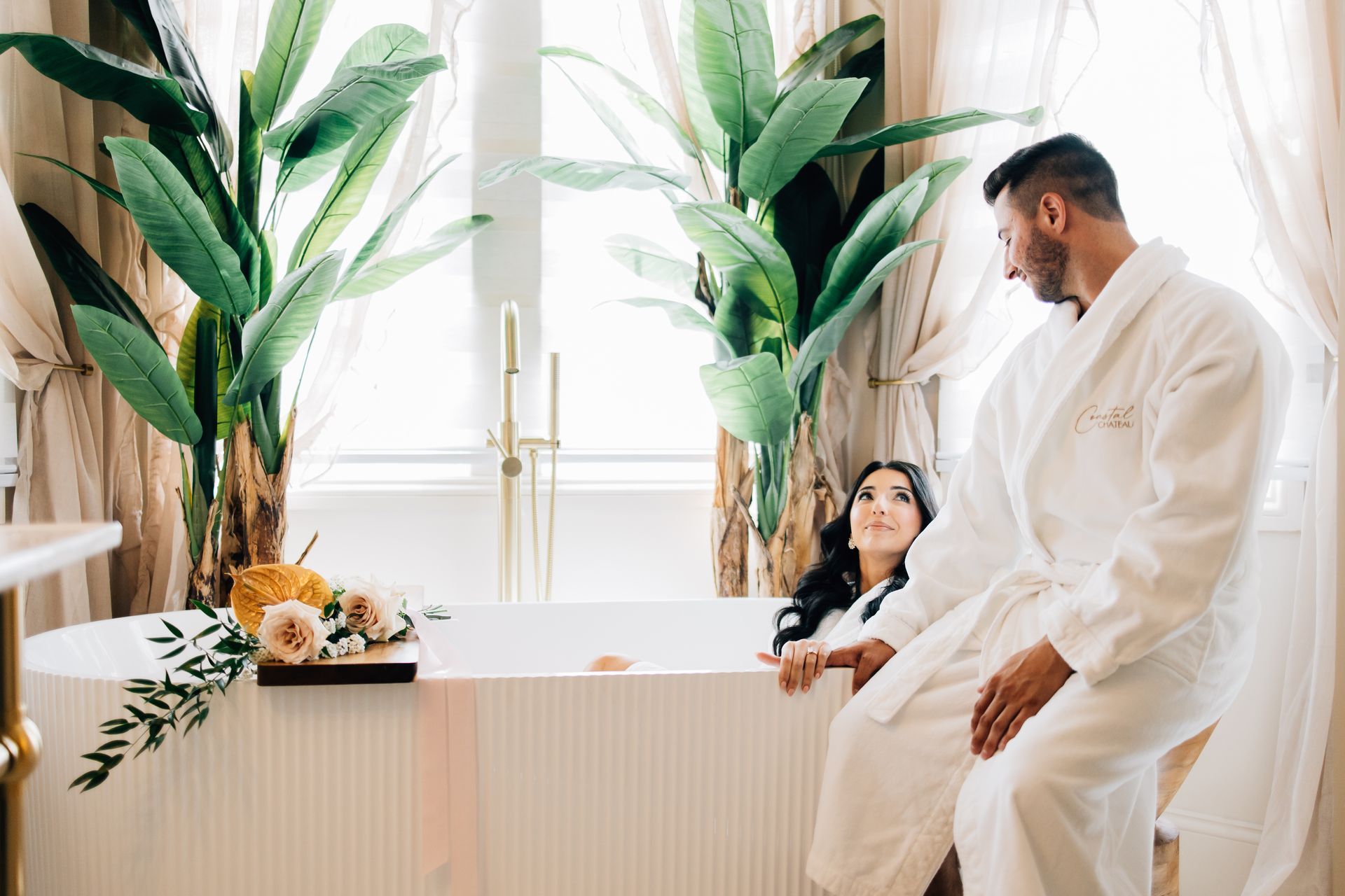 Couple in white robes in a spa-like bathroom, with a bathtub and large plants.
