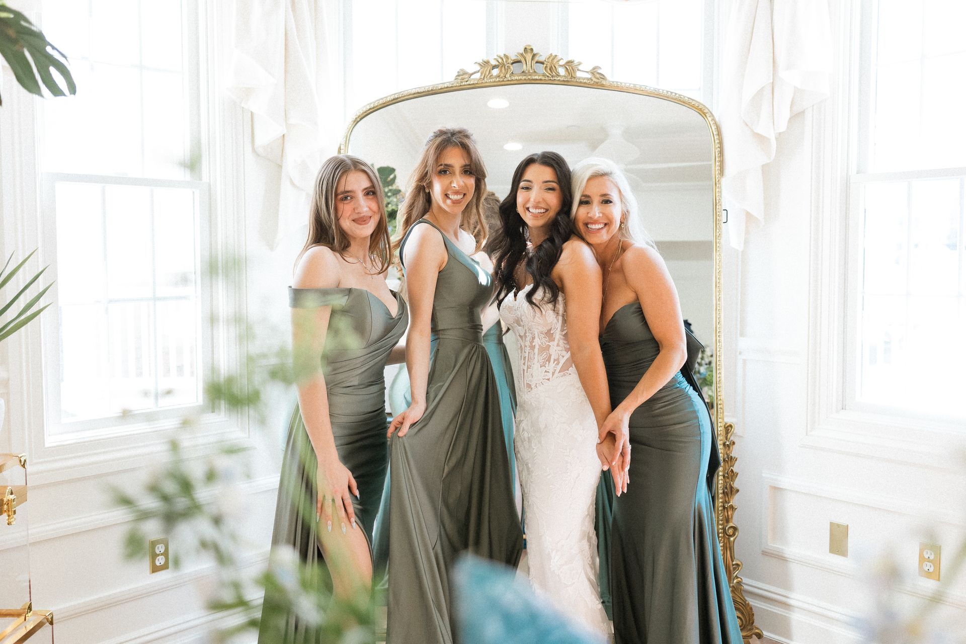 Bride and bridesmaids in dresses posing in front of a mirror in a light-filled room.