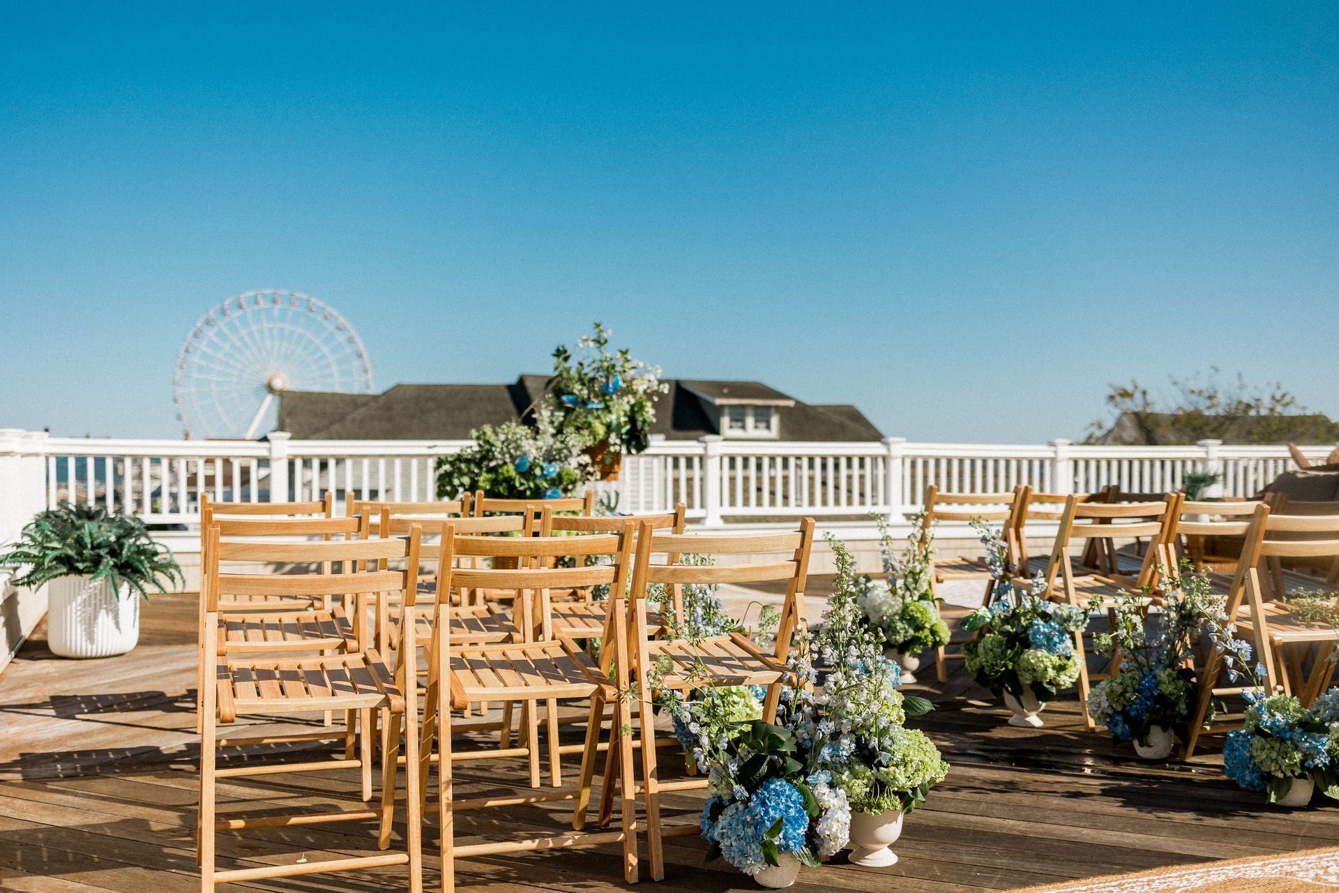 Empty wooden chairs arranged for an outdoor event with blue floral arrangements. Ferris wheel and sky in the background.