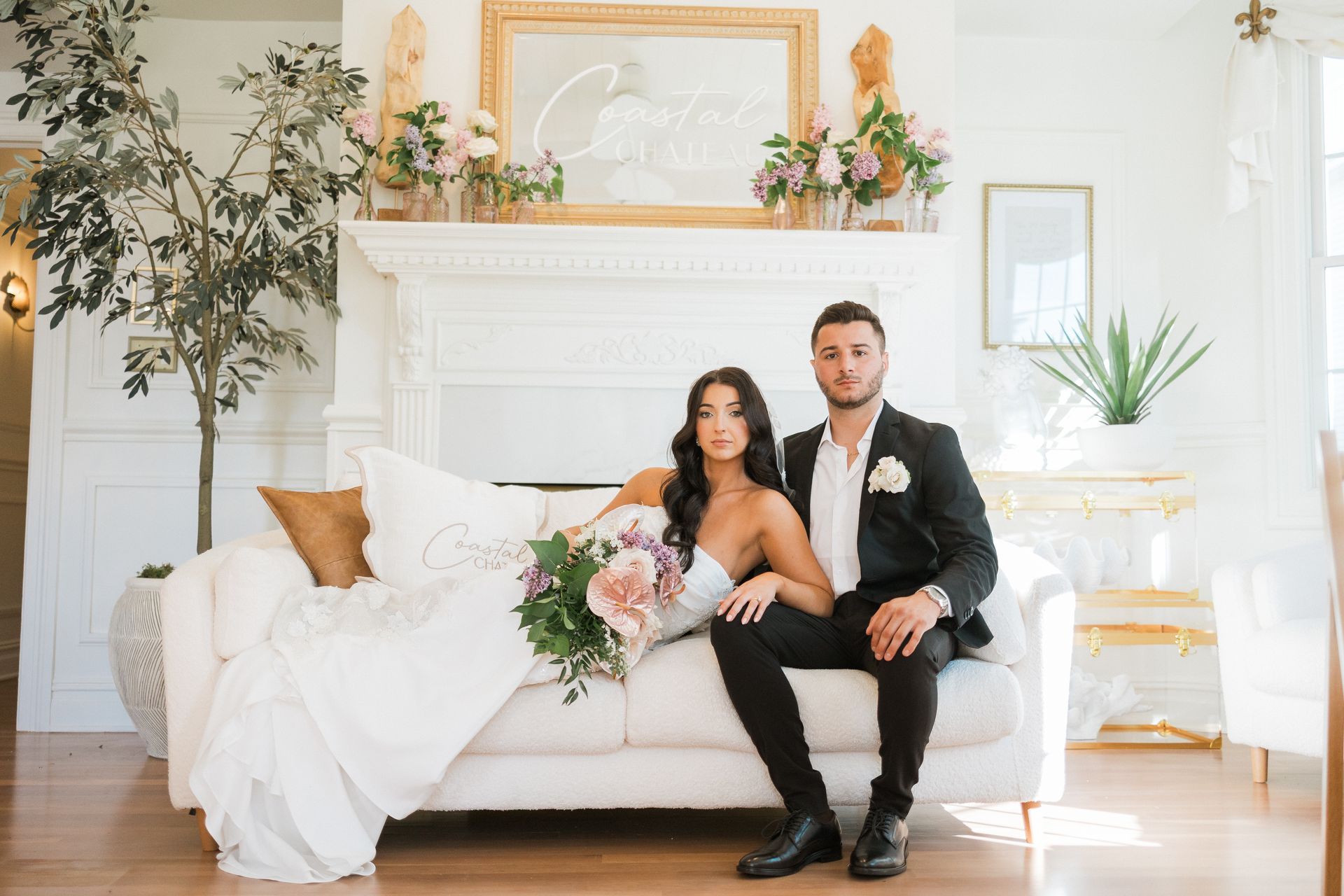 Couple in wedding attire seated on a white couch; smiling in bright room with floral decorations.